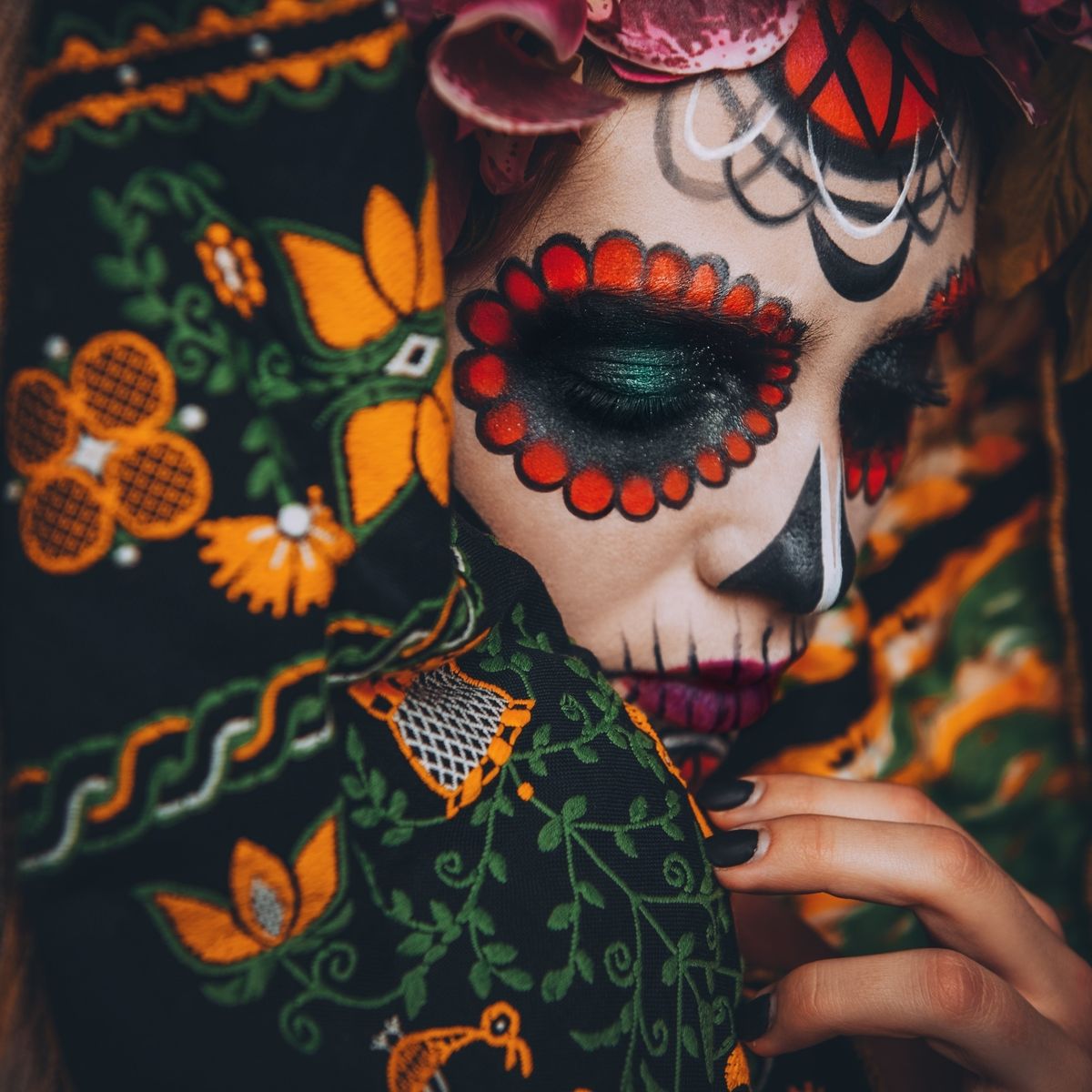 Osoba s barevným líčením na obličeji ve stylu Día di Muertos a zeleným očním stínem, částečně zakrytá zářivou látkou s květinovým vzorem.