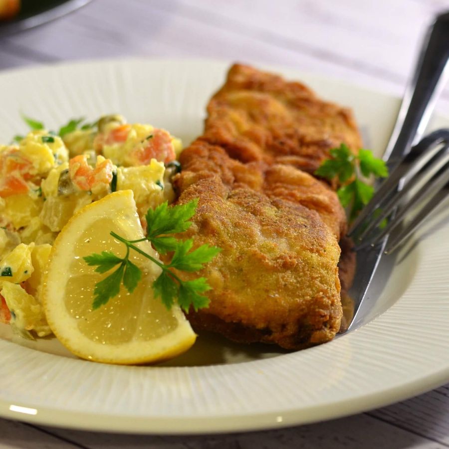 Talíř s obalovaným kaprem, bramborovým salátem zdobeným petrželkou, vidličkou a nožem a plátkem citronu na bílém dřevěném stole.