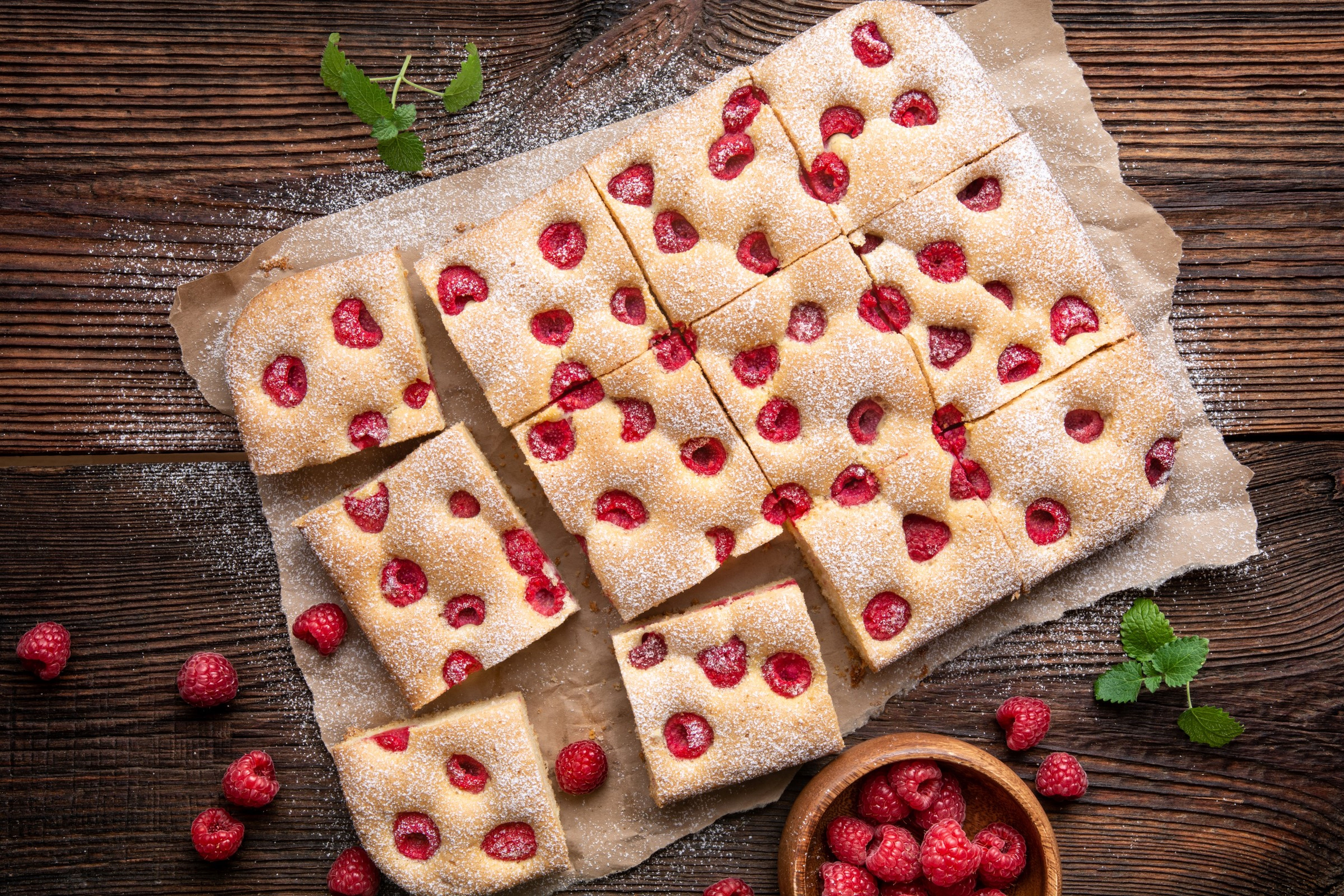 Kousky malinové bublaniny posypané moučkovým cukrem na pečícím papíře, obklopené čerstvými malinami a mátou na dřevěném stole.