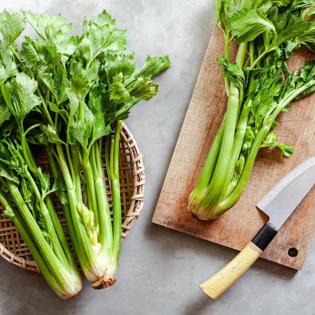 Čerstvé stonky celeru na dřevěné krájecí desce s nožem a v proutěném košíku na šedém povrchu.