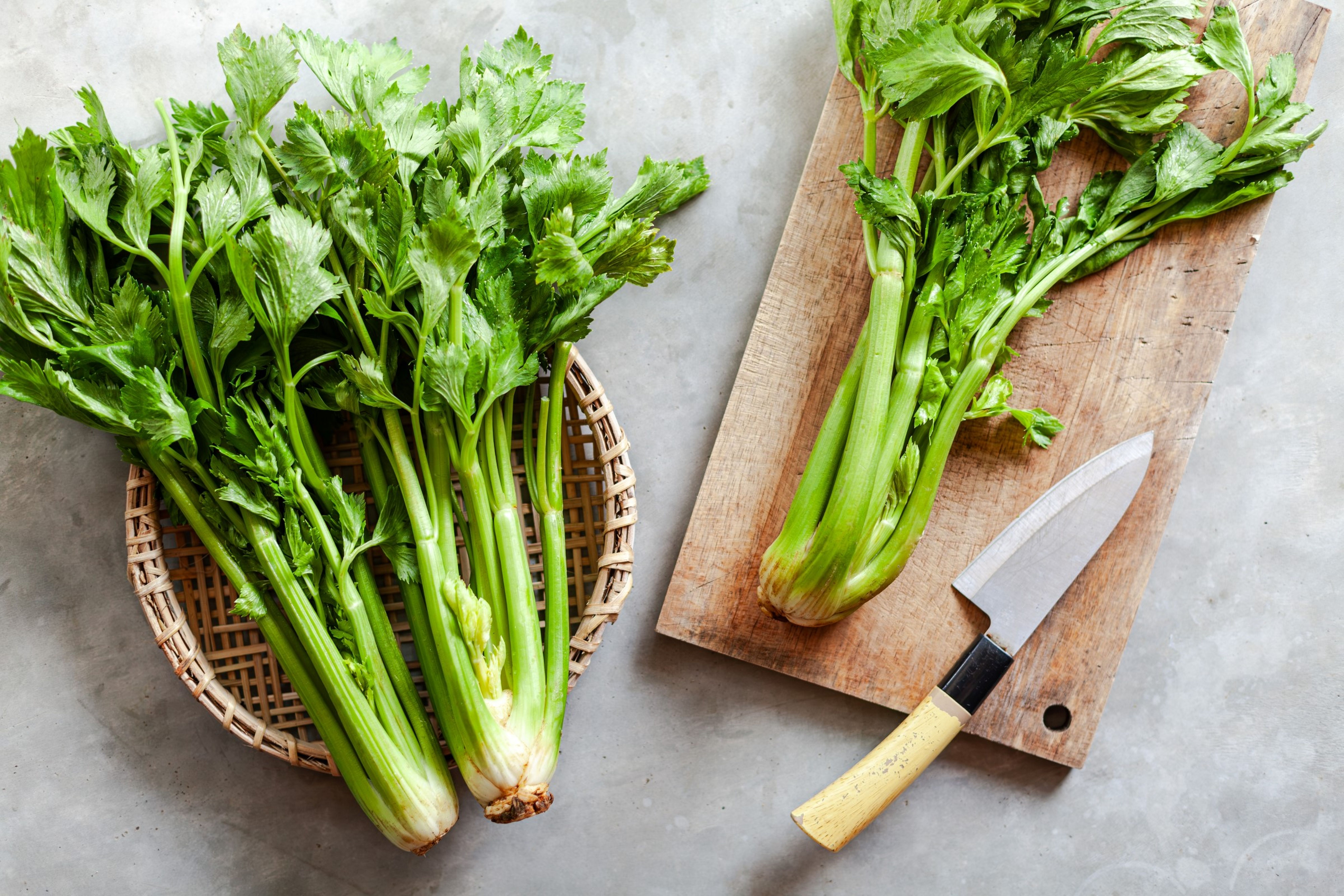 Čerstvé stonky celeru na dřevěné krájecí desce s nožem a v proutěném košíku na šedém povrchu.