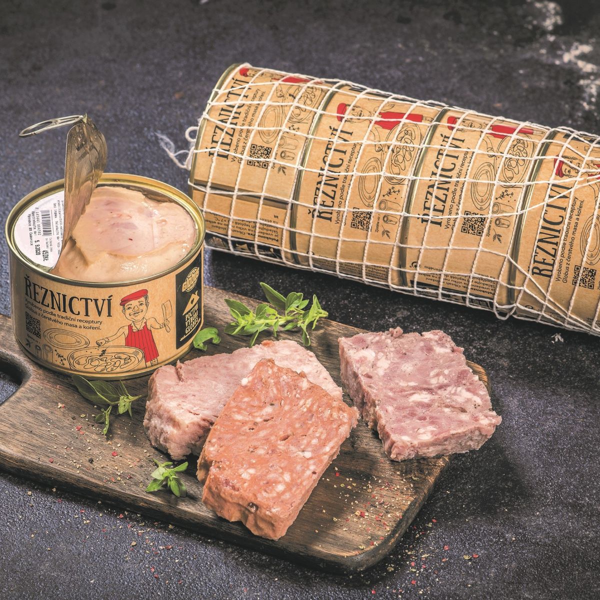 Jedna polokonzerva Globus otevřená, nakrájená různé masové náplně polokonzerv na dřevěné desce s bylinkami, za nimi 4 polokonzervy Globus v síťce, vše položené na tmavém povrchu s texturou.