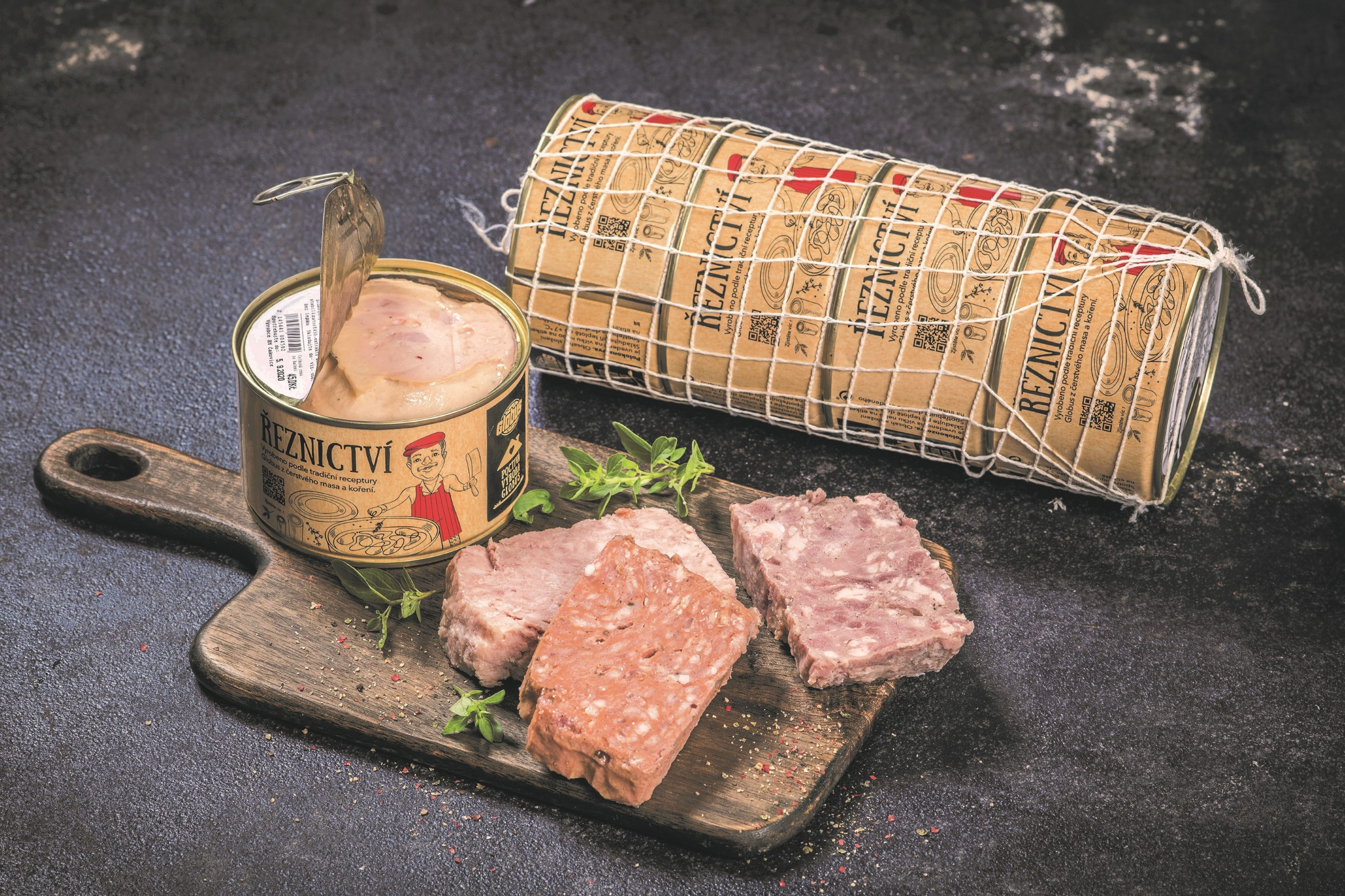 Jedna polokonzerva Globus otevřená, nakrájená různé masové náplně polokonzerv na dřevěné desce s bylinkami, za nimi 4 polokonzervy Globus v síťce, vše položené na tmavém povrchu s texturou.