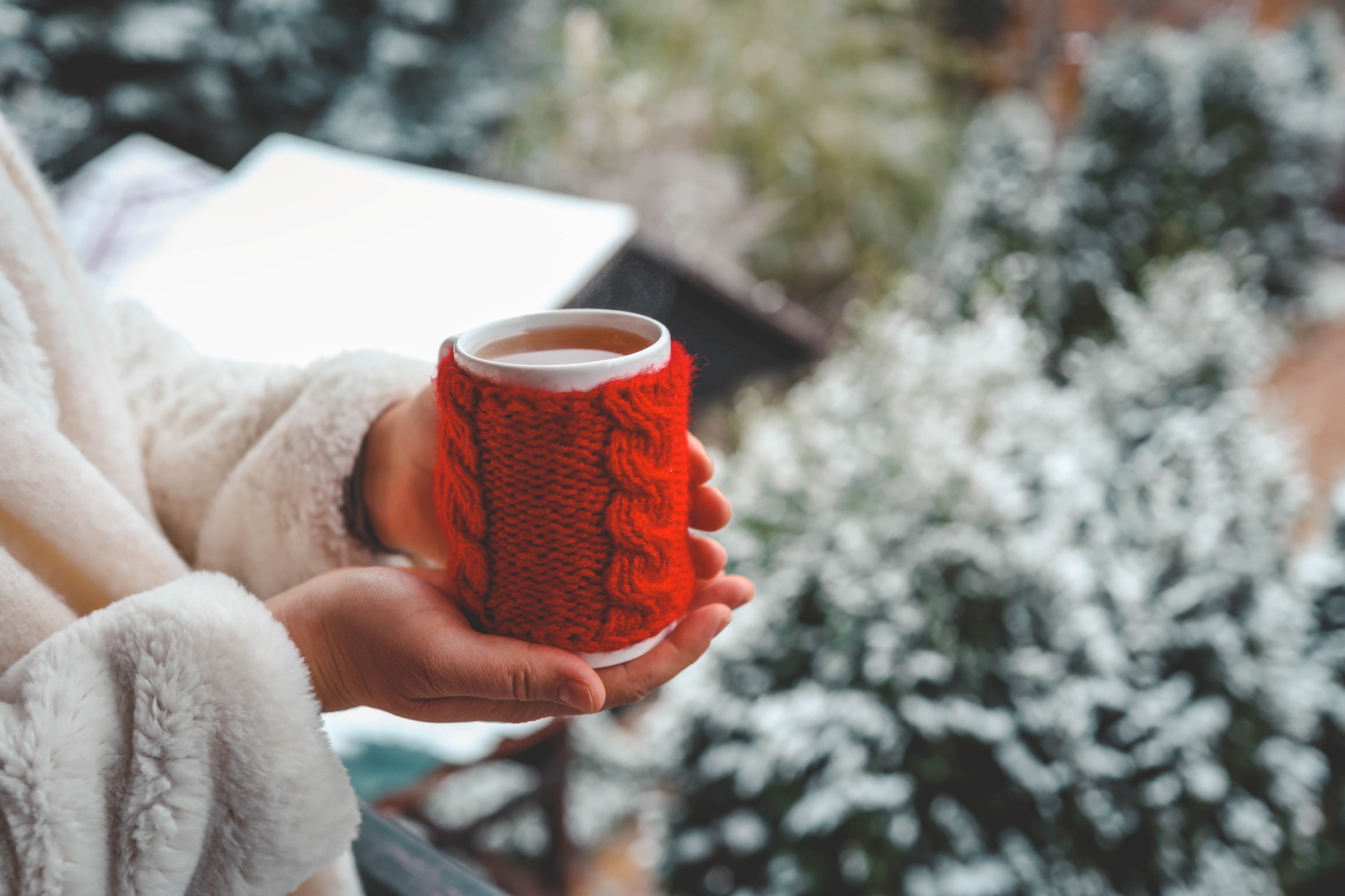 Osoba v bílém kožichu drzí v rukou bílý hrnek s červeným pleteným obalem a v pozadí je vidět zasněžená zahrada.