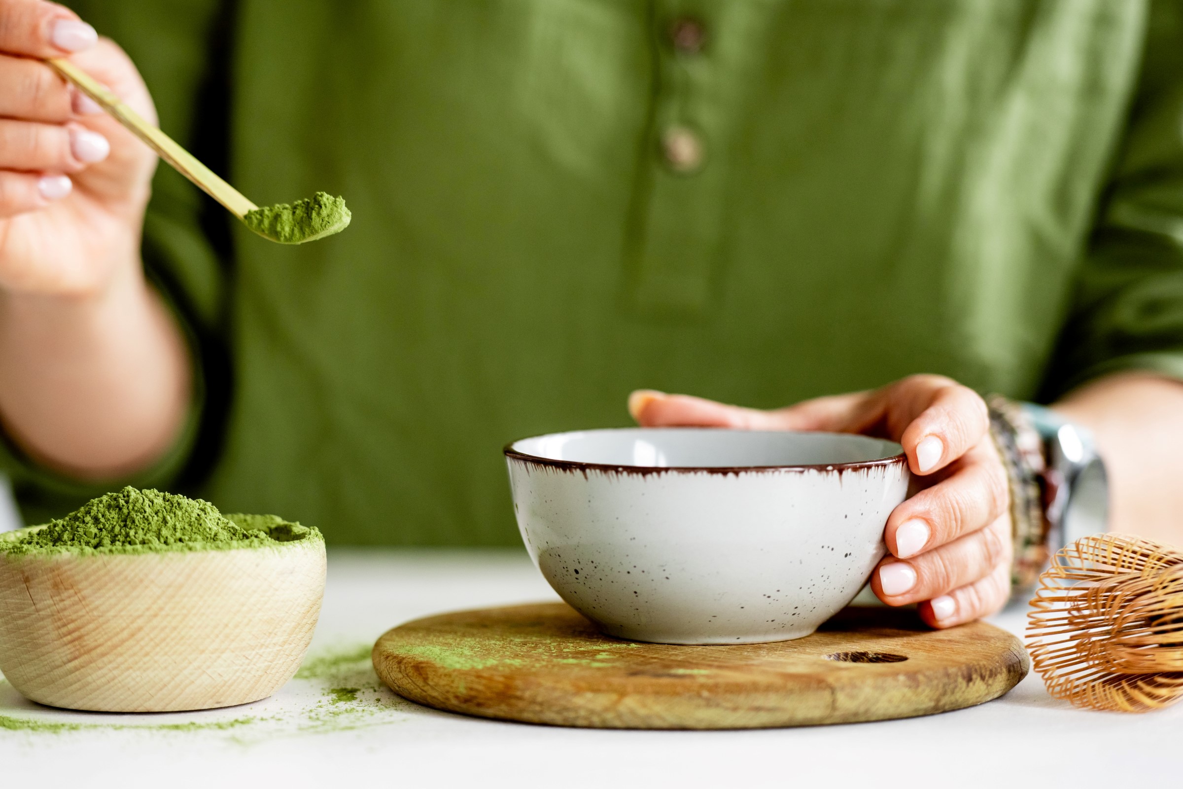 Ruka držící keramickou misku, zatímco nabírá zelený prášek matcha z dřevěné misky; poblíž bambusová metlička.