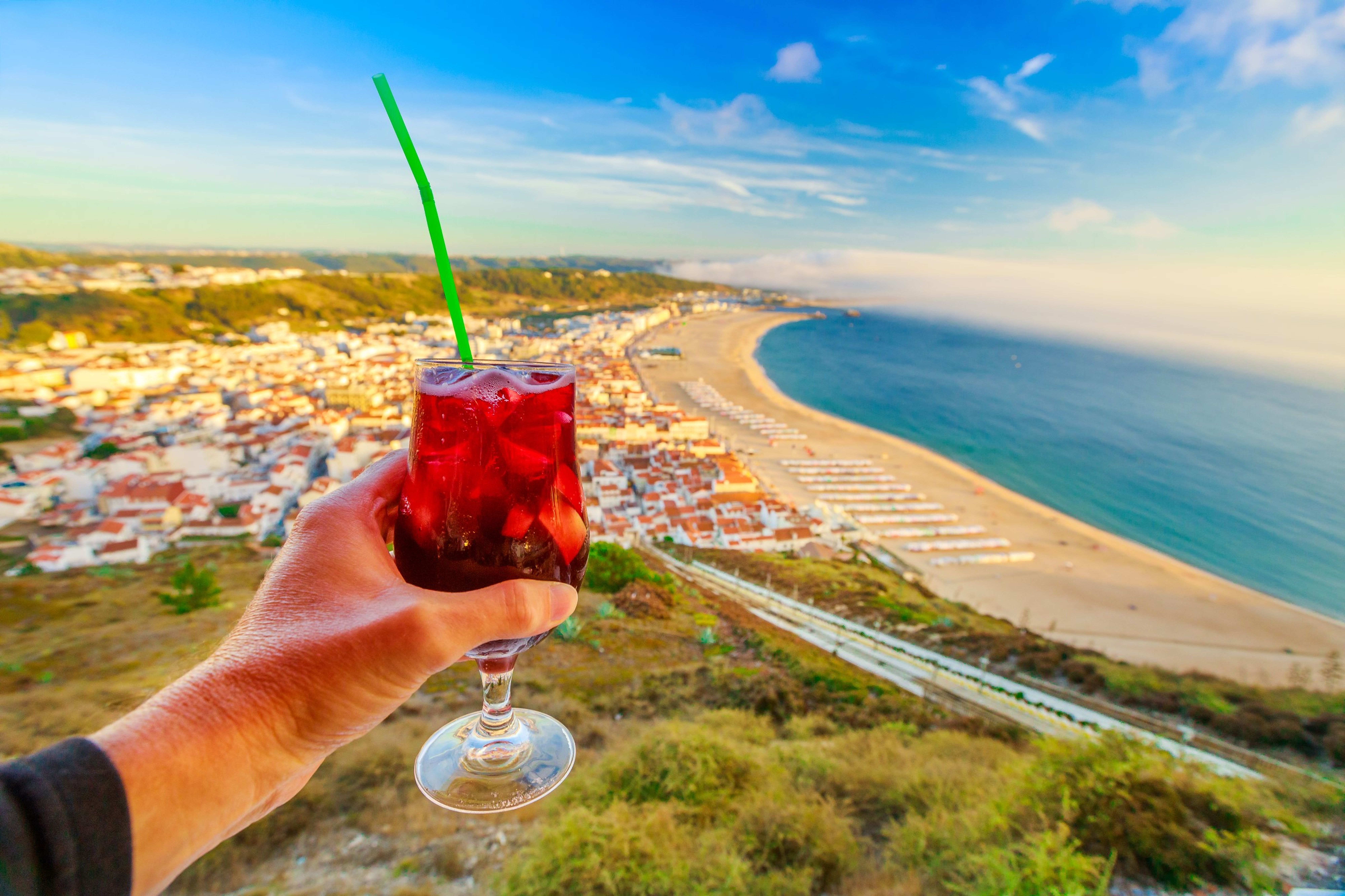 Ruka drží sklenici sangrie se zeleným brčkem a hledí na malebné pobřežní městečko a pláž pod jasně modrou oblohou.