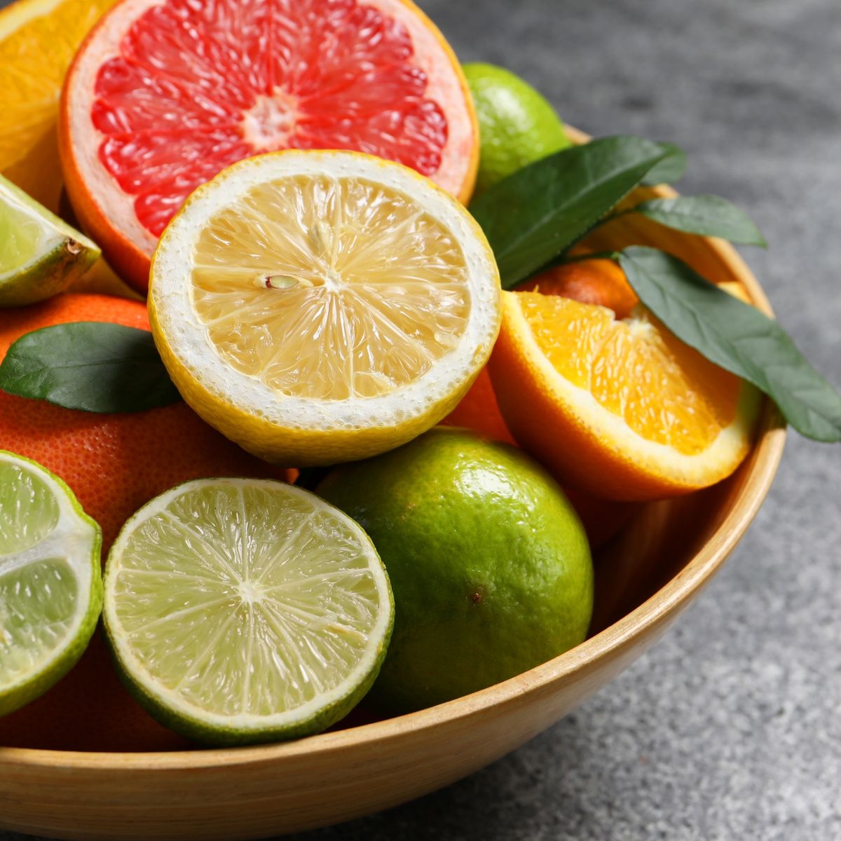 Dřevěná mísa s různými citrusovými plody: rozpůlený grapefruit, citron a limetky, celé pomeranče a zelené listy na šedém kamenném povrchu