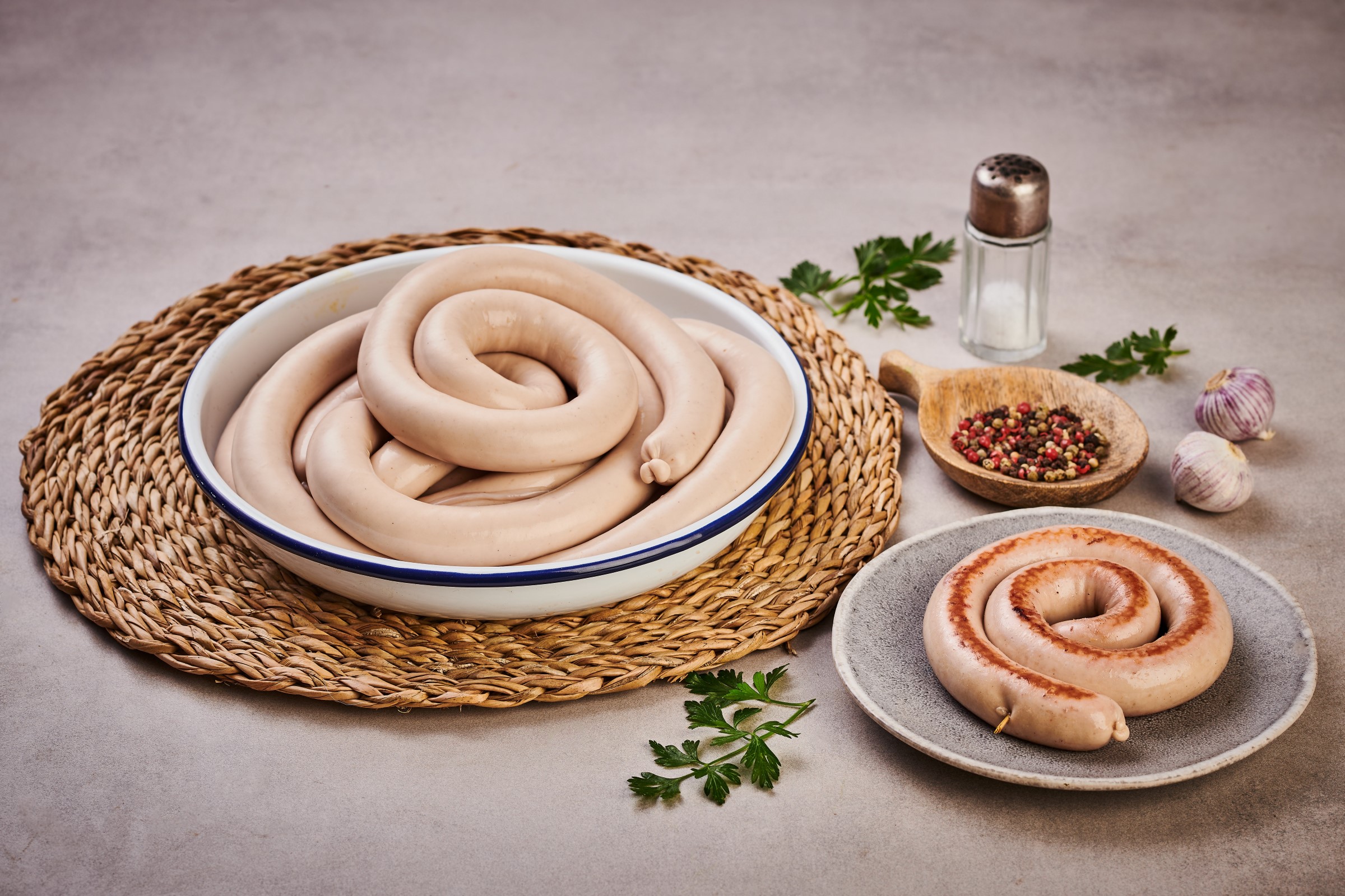 Svinuté vinné klobásy na tkané podložce a talíři, s pepřem, česnekem, petrželkou a solničkou na světlém povrchu.