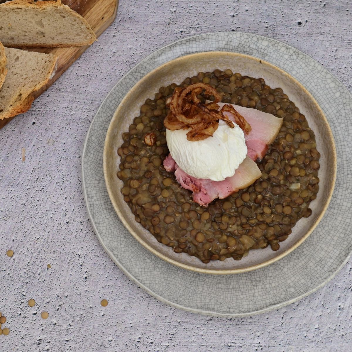Miska čočky na kyselo podávaná s uzeným bokem, tekutým pošírovaným vejcem a křupavou smaženou cibulkou, v pozadí s pečivem. Jedná se o recept z nabídky Globus.