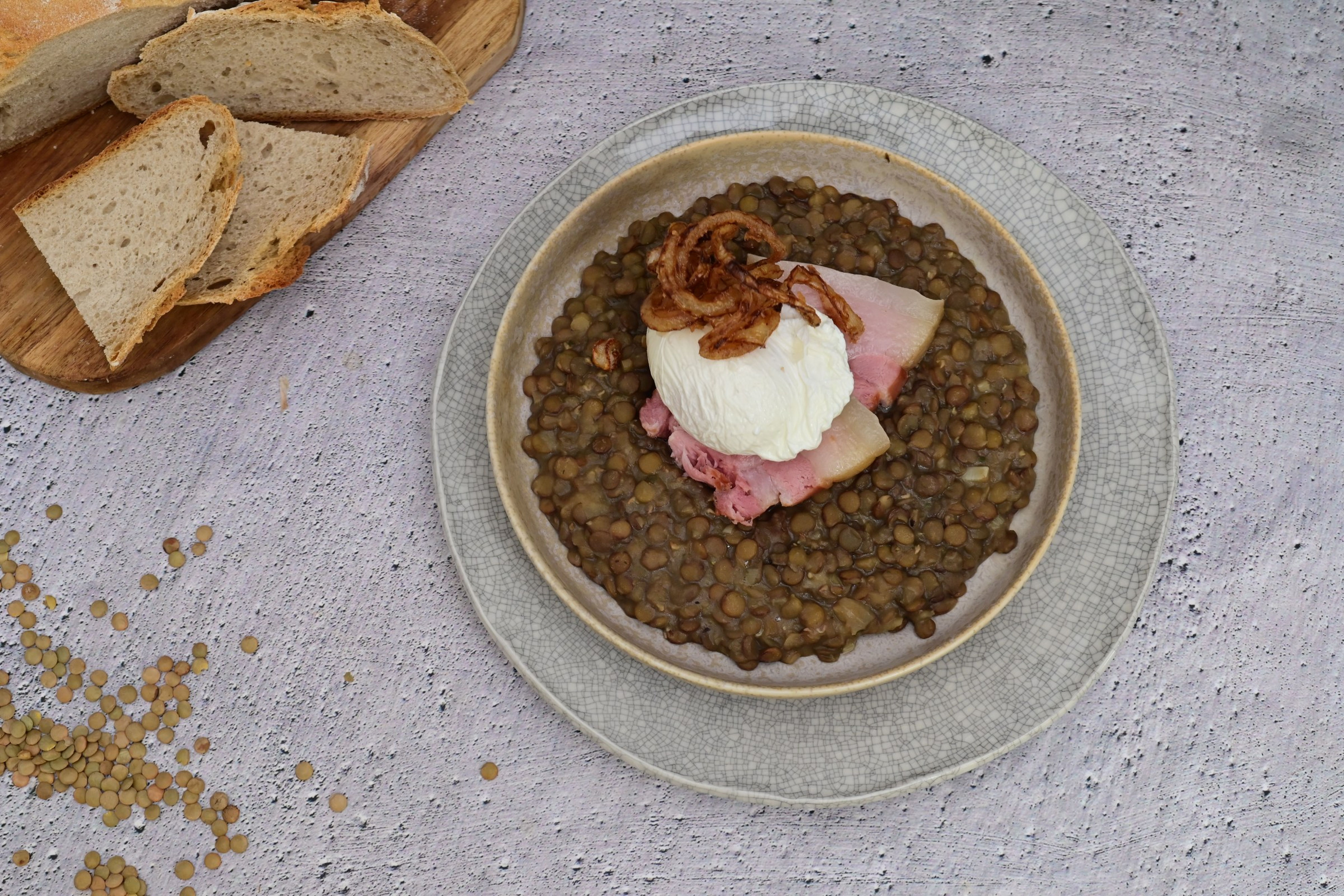 Miska čočky na kyselo podávaná s uzeným bokem, tekutým pošírovaným vejcem a křupavou smaženou cibulkou, v pozadí s pečivem. Jedná se o recept z nabídky Globus.