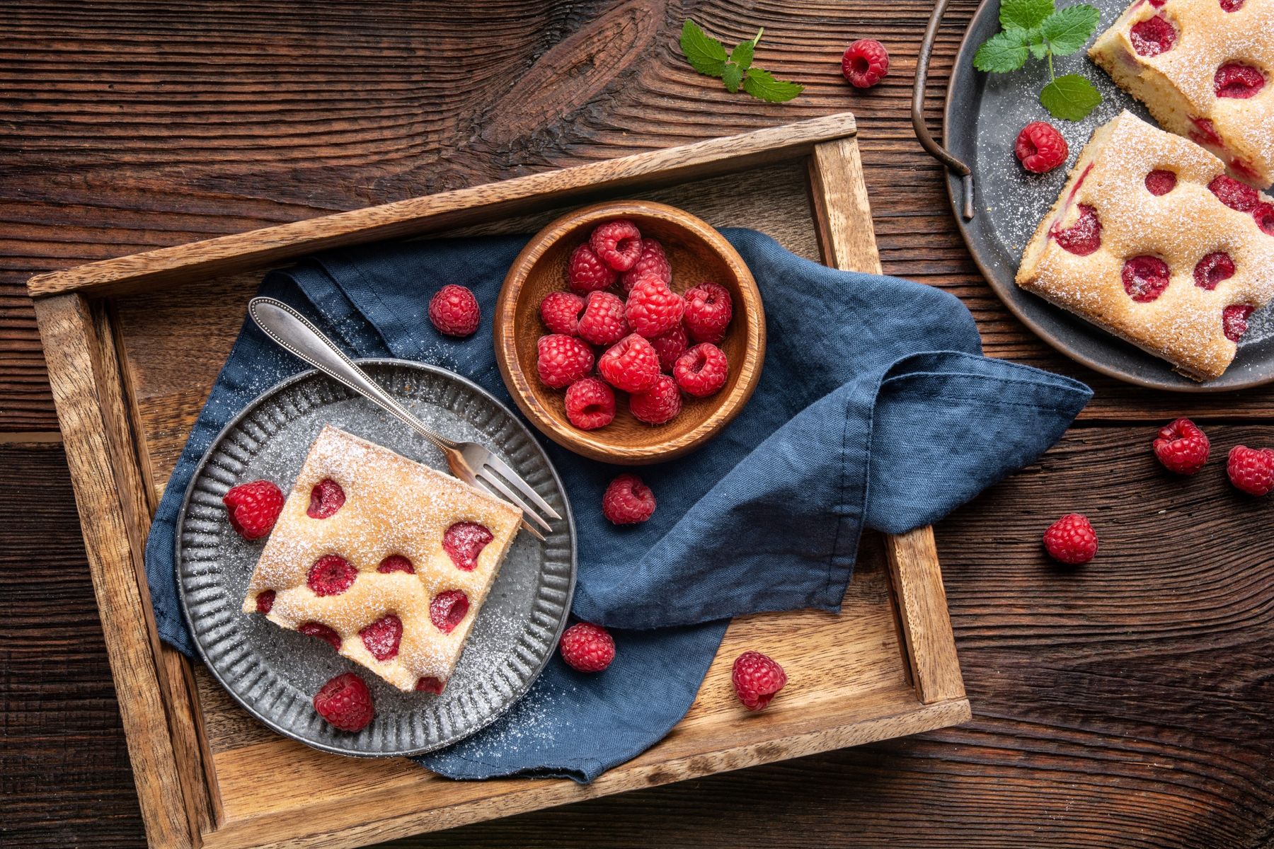 Dřevěný podnos s kousky malinové bublaniny, mísa s čerstvými malinami, stříbrná vidlička a modrý ubrus na dřevěném stole.
