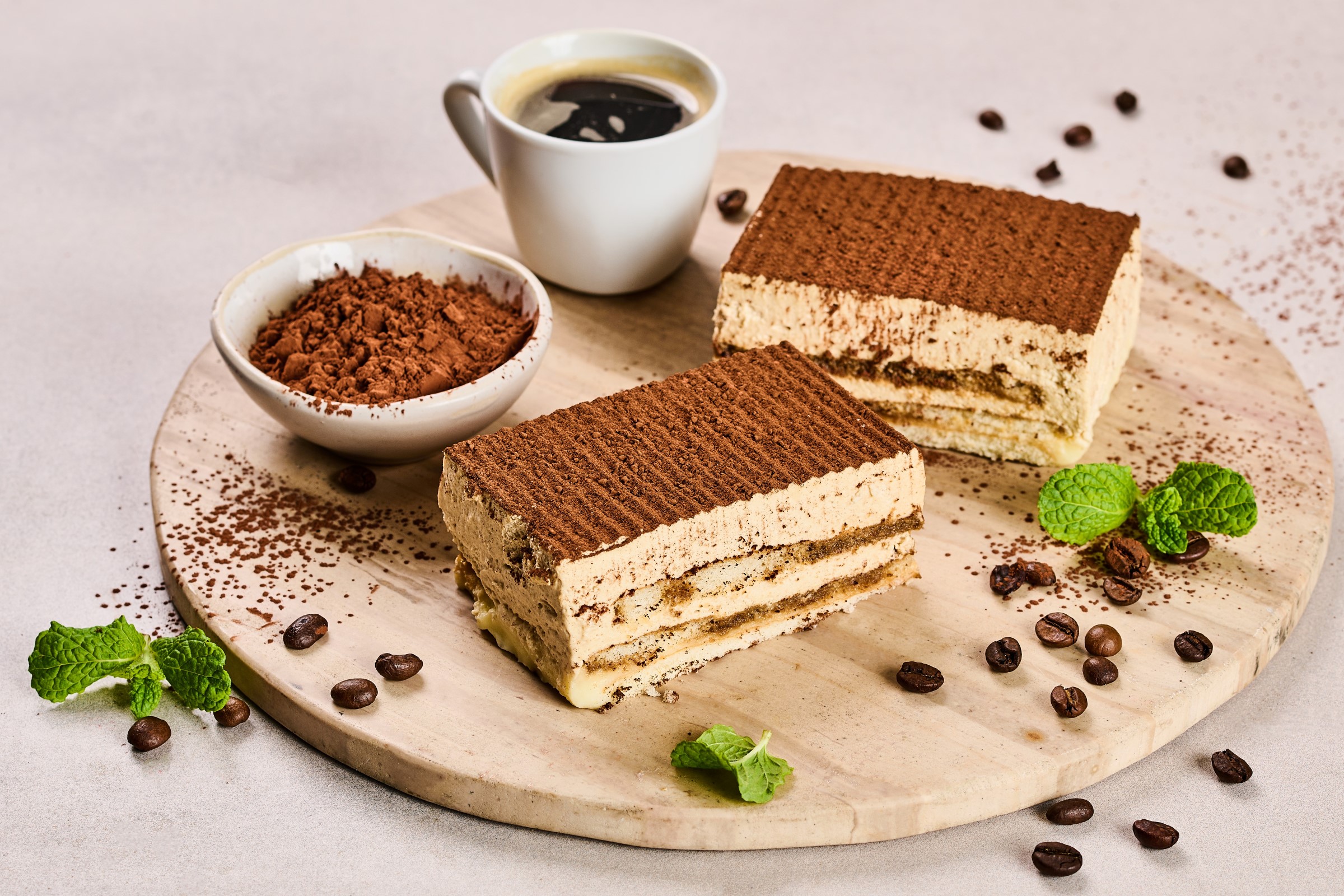 Dva kousky řezu tiramisu na dřevěné podložce, k nim šálek kávy, kakaový prášek, kávová zrna a lístky čerstvé máty.