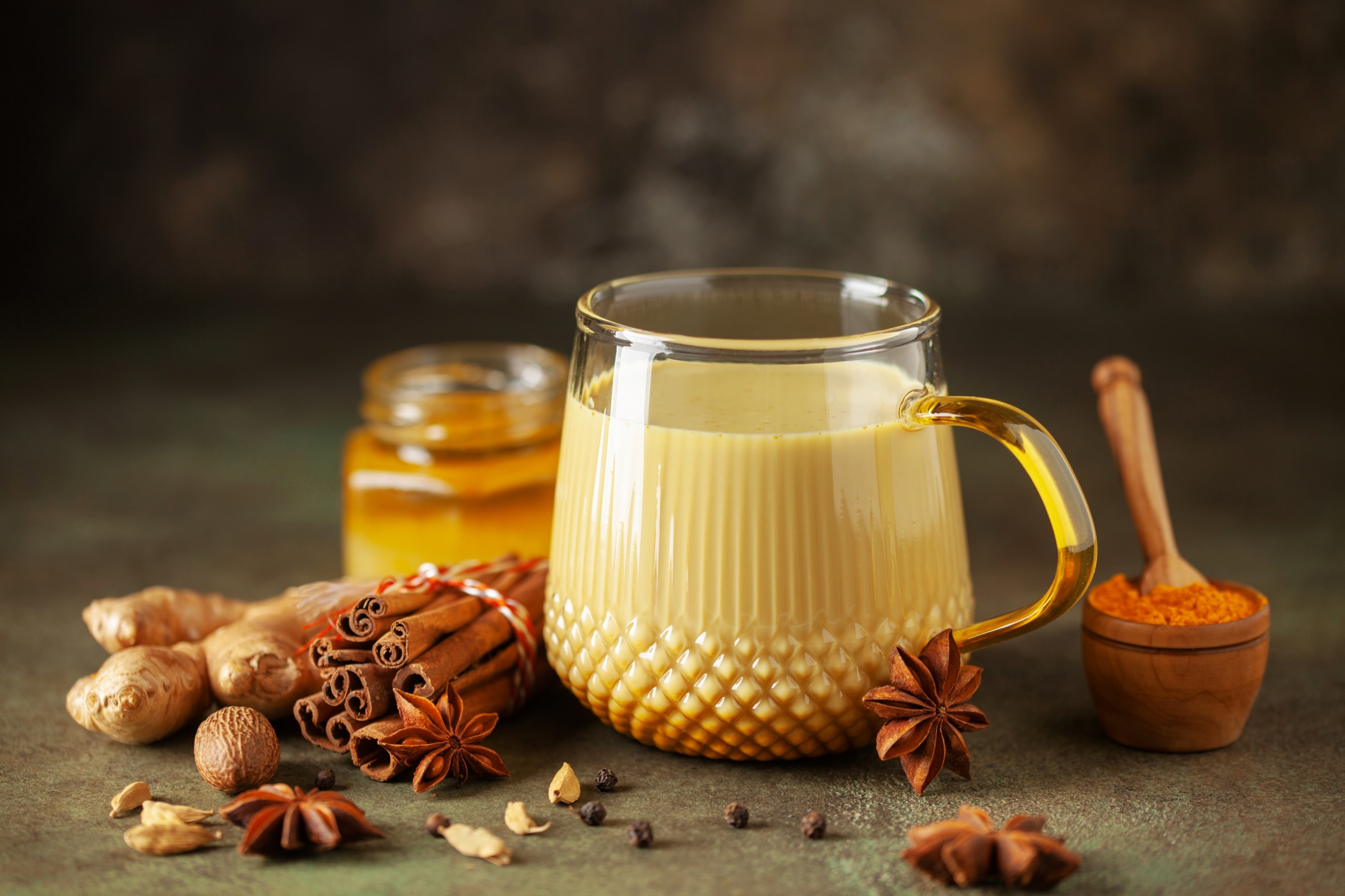 Skleněný hrnek se zlatým kurkumovým mlékem obklopený sklenicí medu, tyčinkami skořice, zázvorem, badyánem, muškátovým oříškem a kurkumovým práškem.