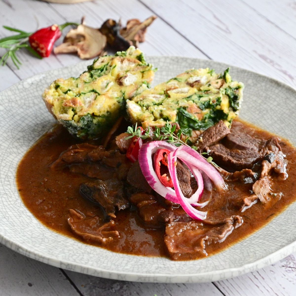 Talíř se srnčím masem v husté omáčce, ozdobený červenou cibulí a chilli, doplněný dvěma plátky špenátového knedlíku. Recept připravil Vojta Poštulka.