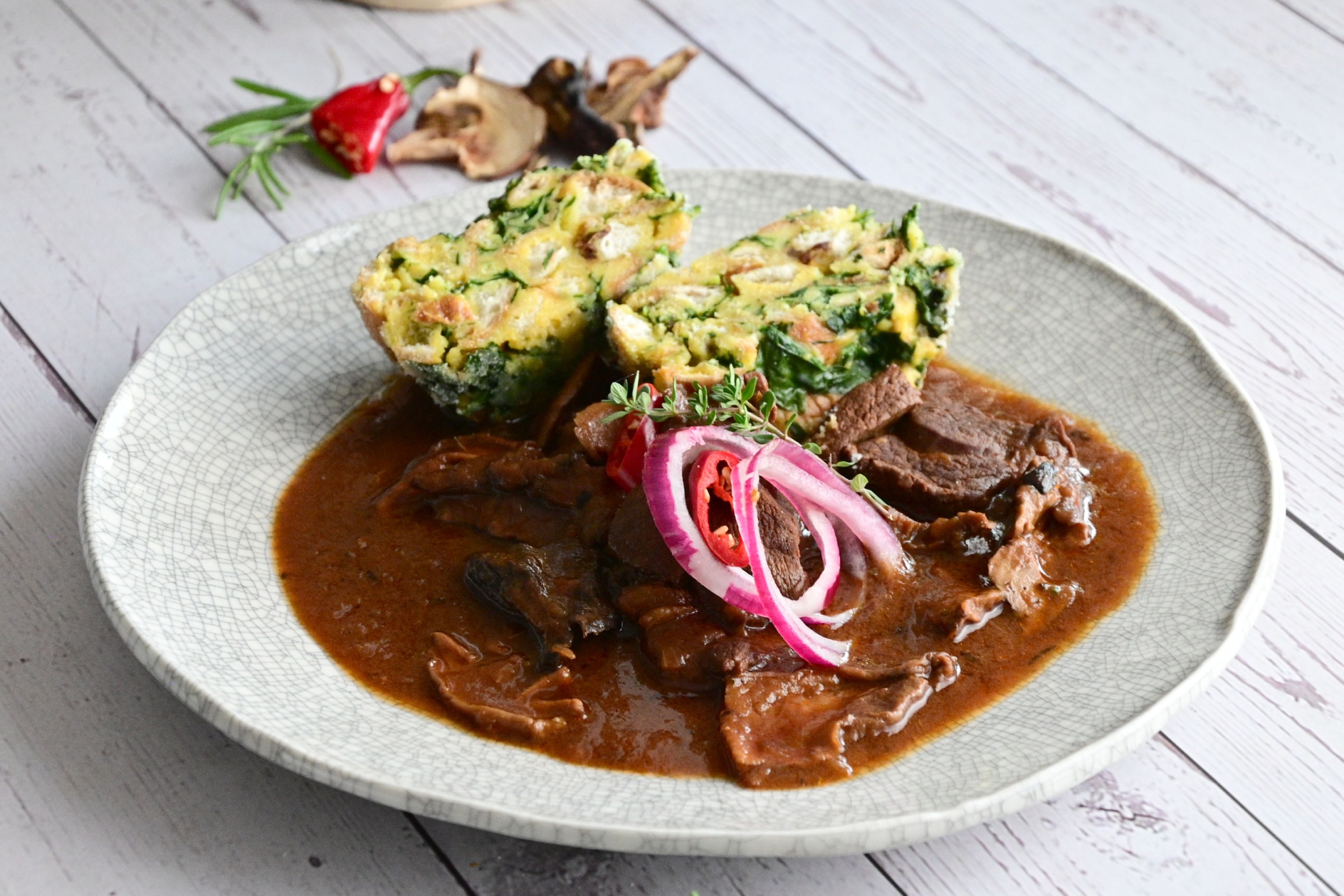 Talíř se srnčím masem v husté omáčce, ozdobený červenou cibulí a chilli, doplněný dvěma plátky špenátového knedlíku. Recept připravil Vojta Poštulka.