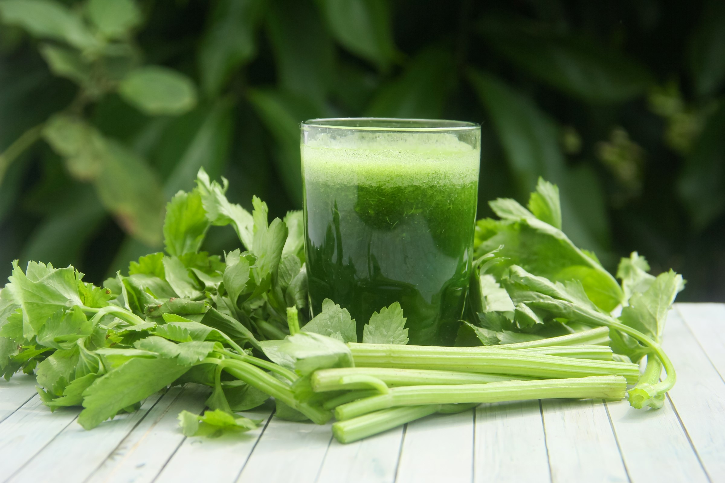 Sklenice zelené šťávy z řapíkatého celeru obklopená čerstvými stonky celeru na dřevěné ploše, s bujnými zelenými listy v pozadí.