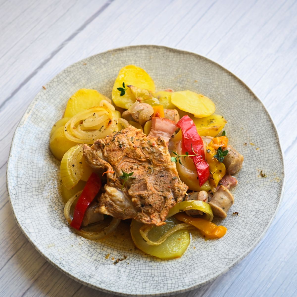 Jídlo s pečeným masem, nakrájenými bramborami, cibulí, paprikou a bylinkami na rustikálním strukturovaném talíři. Recept připravil Vojta Poštulka.