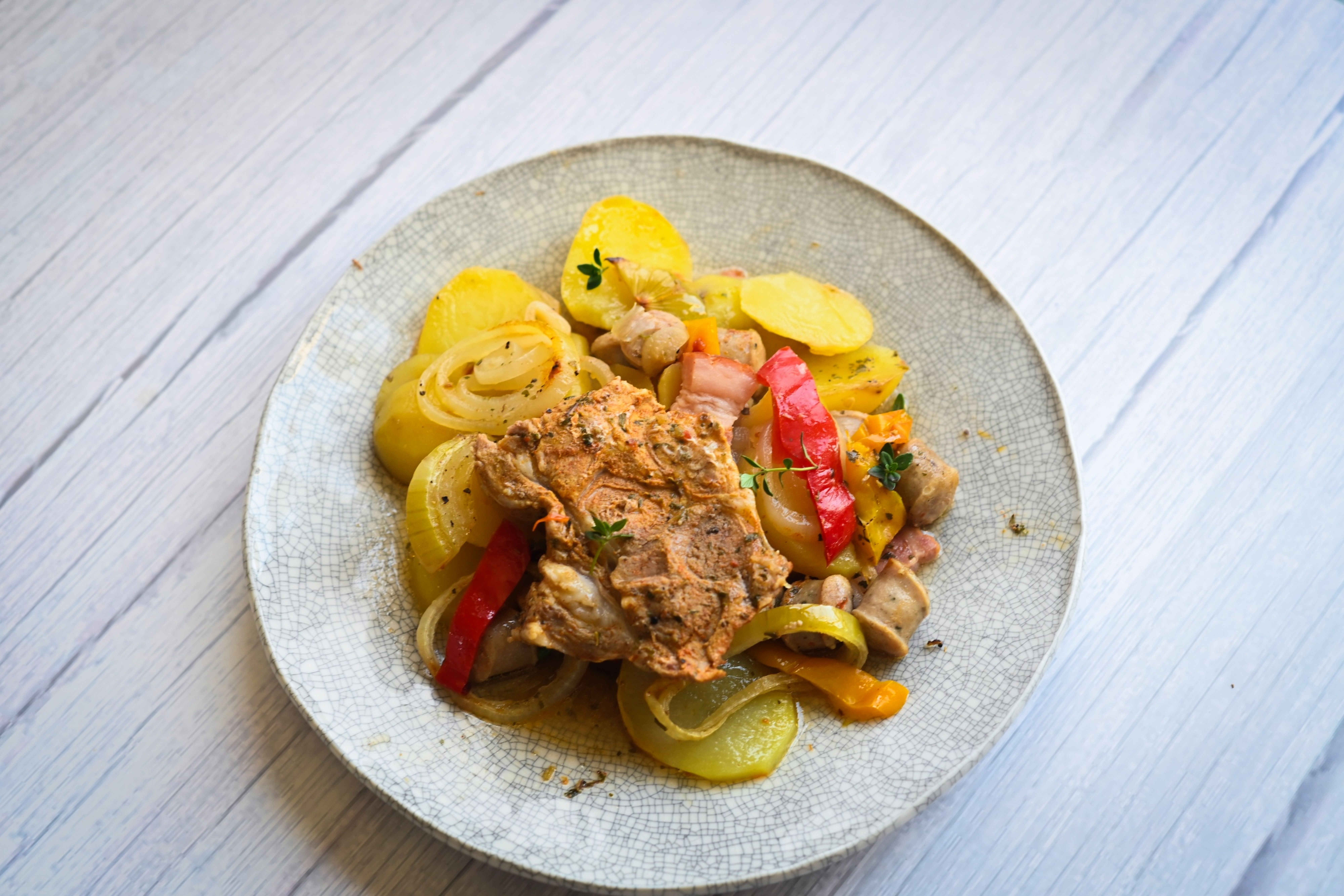 Jídlo s pečeným masem, nakrájenými bramborami, cibulí, paprikou a bylinkami na rustikálním strukturovaném talíři. Recept připravil Vojta Poštulka.