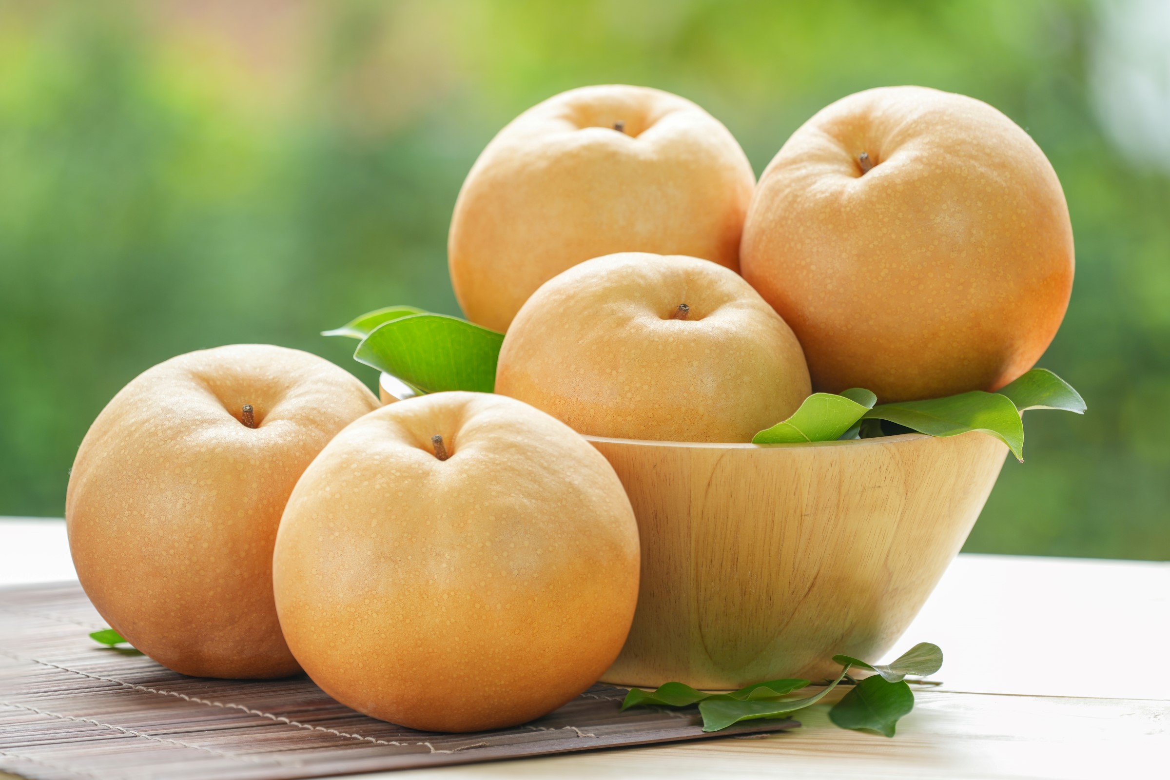 Dřevěná mísa naplněná pěti čerstvými asijskými hruškami nashi, obklopená zelenými listy, umístěná na bambusové rohoži s rozmazaným zeleným pozadím.