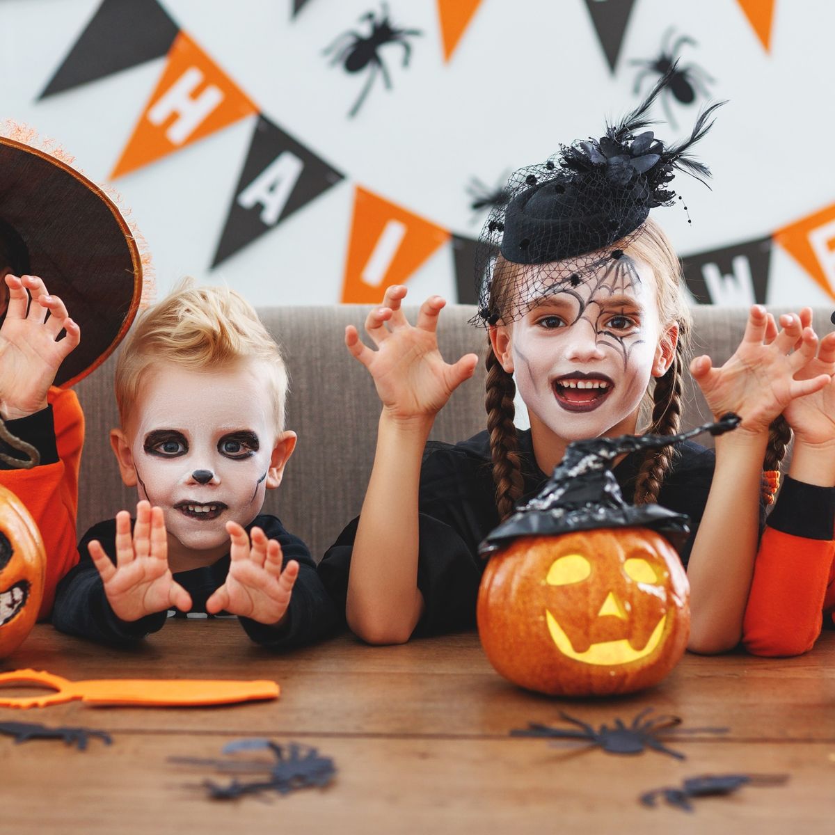 Děti v halloweenských kostýmech s namalovanými obličeji, které mají hravé výrazy, obklopené vyřezanými dýněmi na stole, s halloweenskou výzdobou.