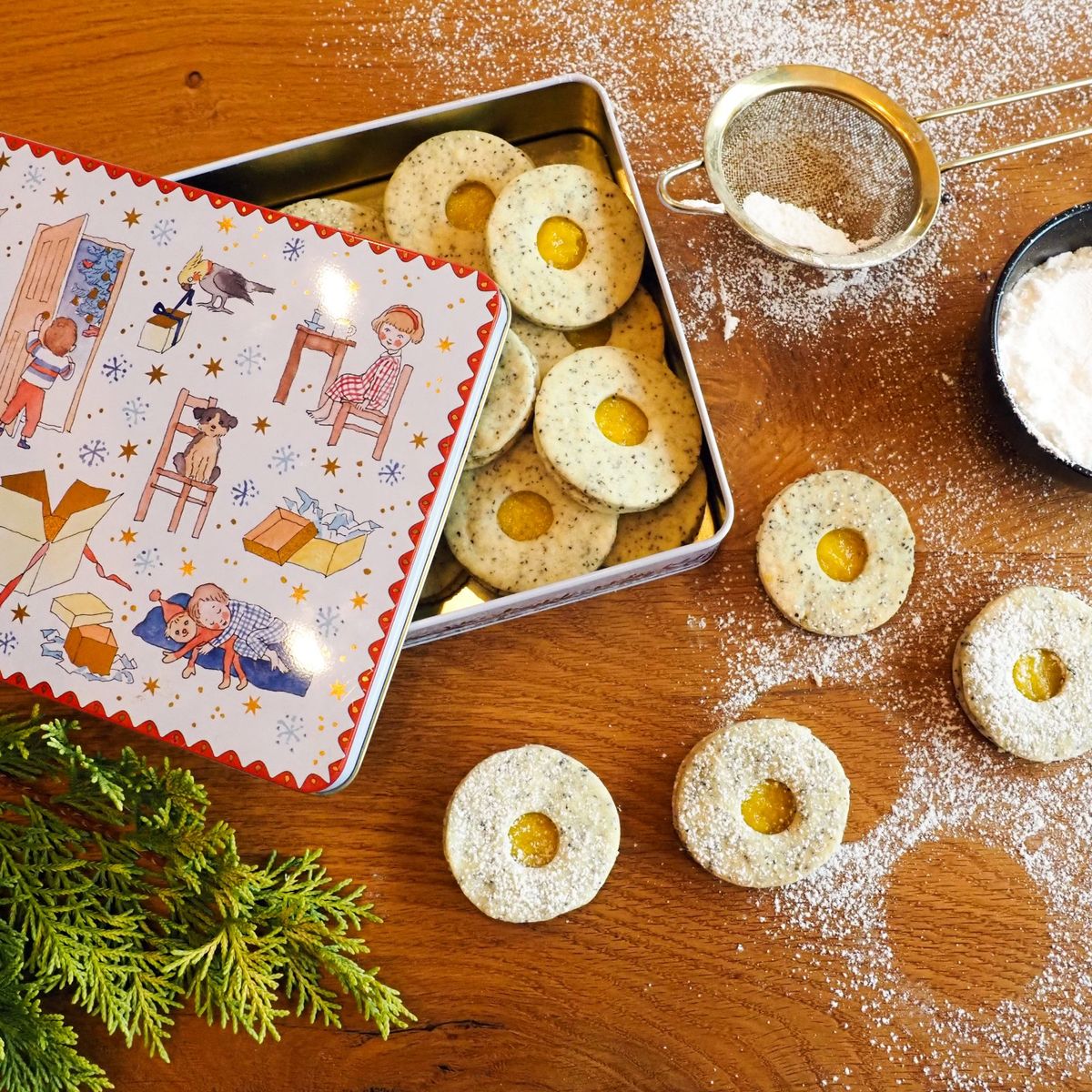 Dóza plná makových lineckých, sítko, moučkový cukr a větvičky jehličí na dřevěném stole, podle receptu na veganské linecké cukroví od Evy z Kitchen Affair z webu hypermarketu Globus.