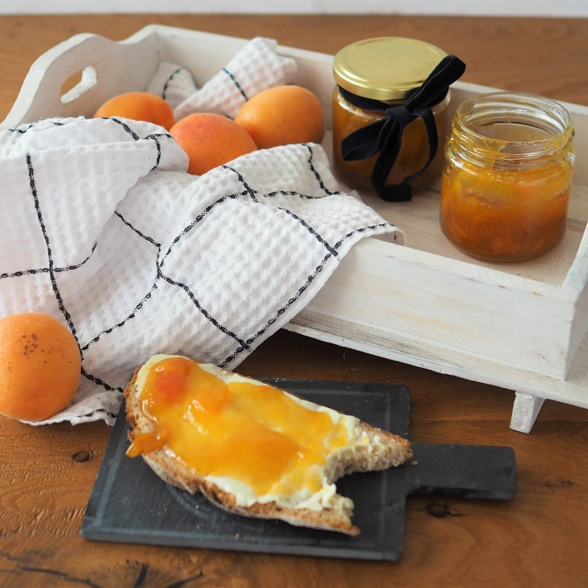 Meruňky na bílém podnosu spolu se sklenicemi meruňkové marmelády, bílým ubrusem a toastem s marmeládou na malé černé desce.