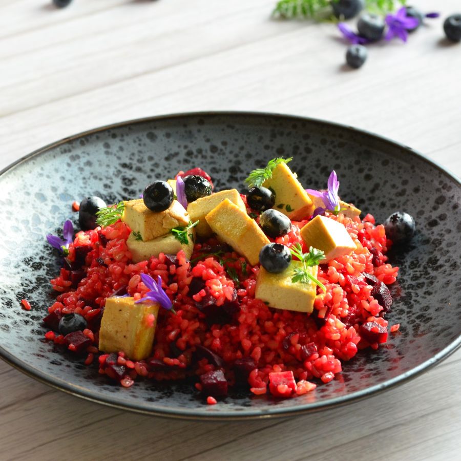 Černá miska s jasně červenou rýží posypaná kostičkami tofu, borůvkami, červenou řepou, bylinkami a fialovými jedlými květy.