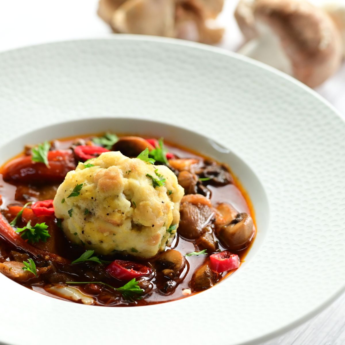 Miska houbového guláše se zeleninou a kefírovým knedlíkem, ozdobená bylinkami a plátky červené chilli papričky, vedle hrnec s gulášem na světlém dřevěném povrchu, podle receptu z hypermarketu Globus od Vojty Poštulky.