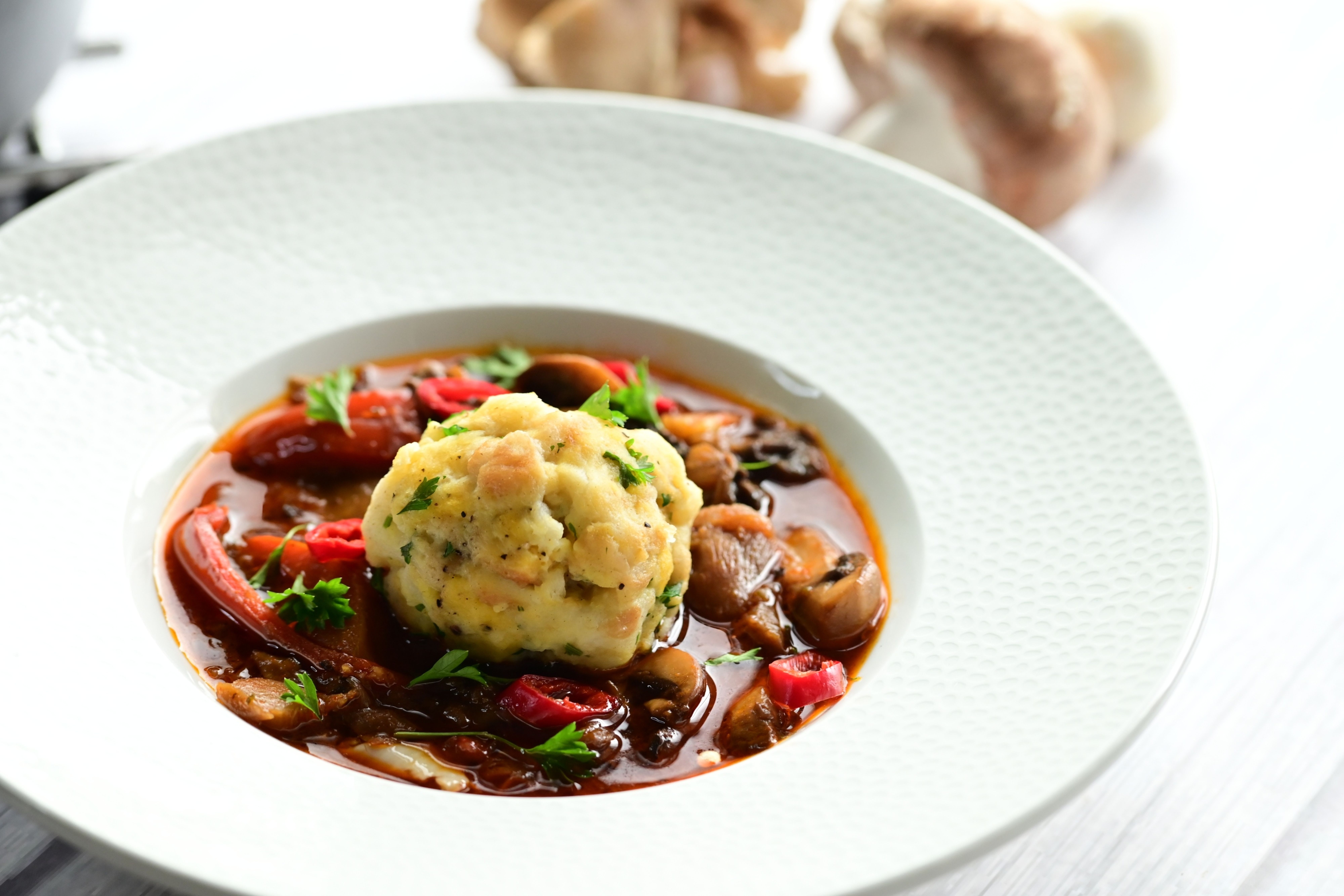 Miska houbového guláše se zeleninou a kefírovým knedlíkem, ozdobená bylinkami a plátky červené chilli papričky, vedle hrnec s gulášem na světlém dřevěném povrchu, podle receptu z hypermarketu Globus od Vojty Poštulky.