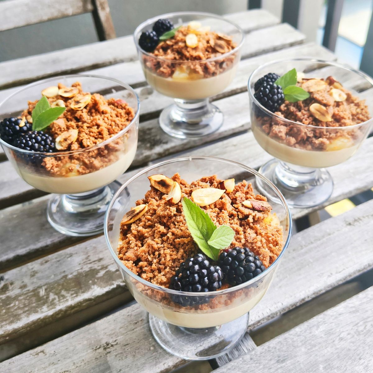 Čtyři skleničky s krémovým základem, ozdobené drobenkou, ostružinami, plátky mandlí a lístky máty na rustikálním dřevěném stole. Recept připravily Ségry v kuchyni.