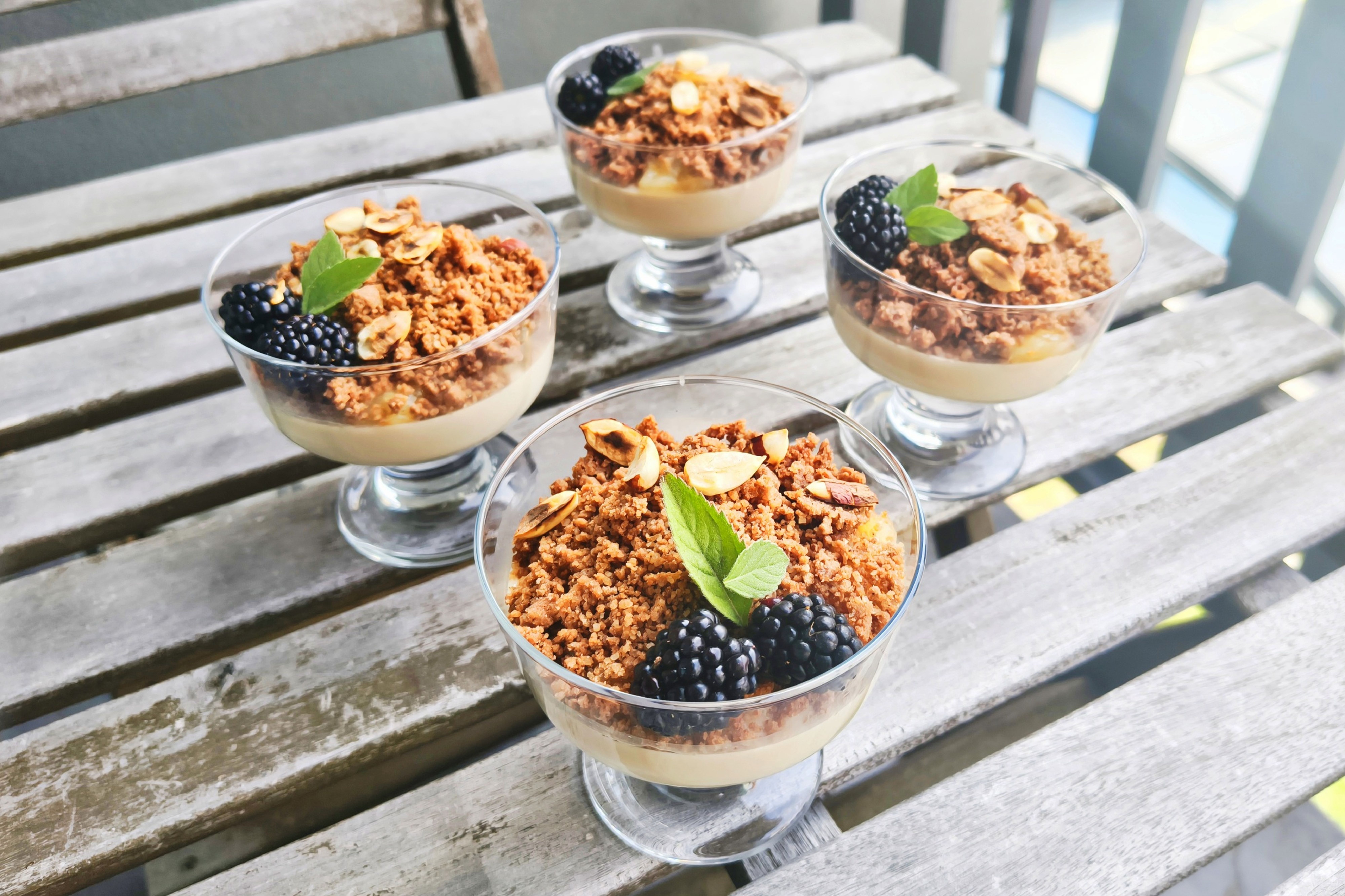 Čtyři skleničky s krémovým základem, ozdobené drobenkou, ostružinami, plátky mandlí a lístky máty na rustikálním dřevěném stole. Recept připravily Ségry v kuchyni.