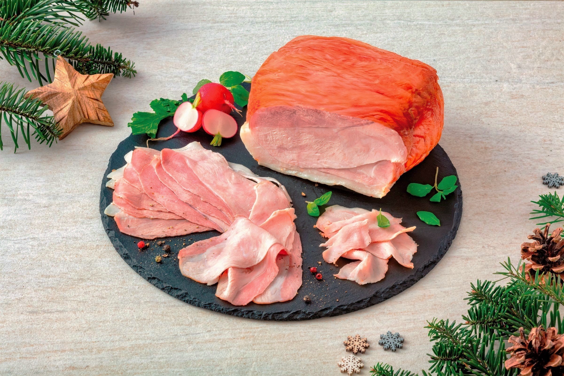 Nakrájená Vánoční šunka Globus na kulatém břidlicovém talíři s ředkvičkami a bylinkami, obklopená slavnostními borovicovými větvičkami a dřevěnou hvězdou.