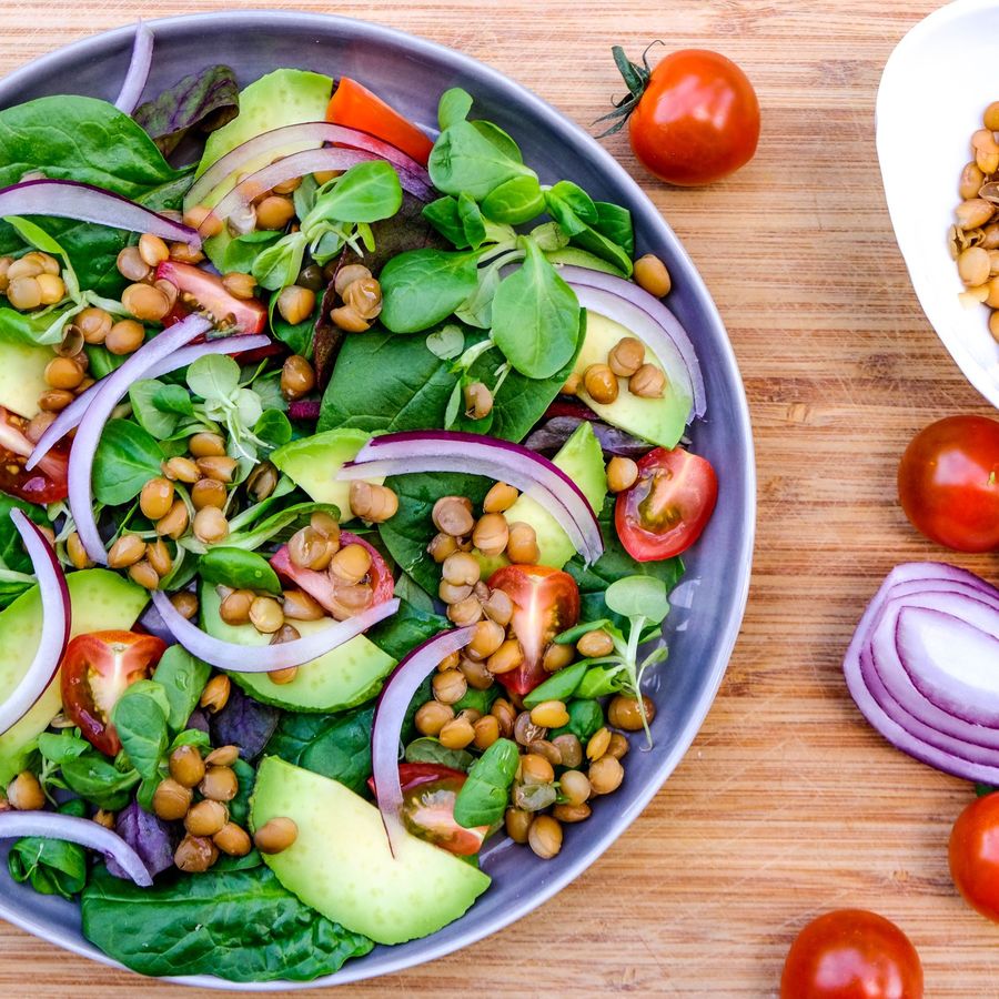 Směs s listovým salátem, avokádem, červenou cibulí, cherry rajčátky a čočkou v misce. Ingredience jsou také rozloženy na dřevěné desce.