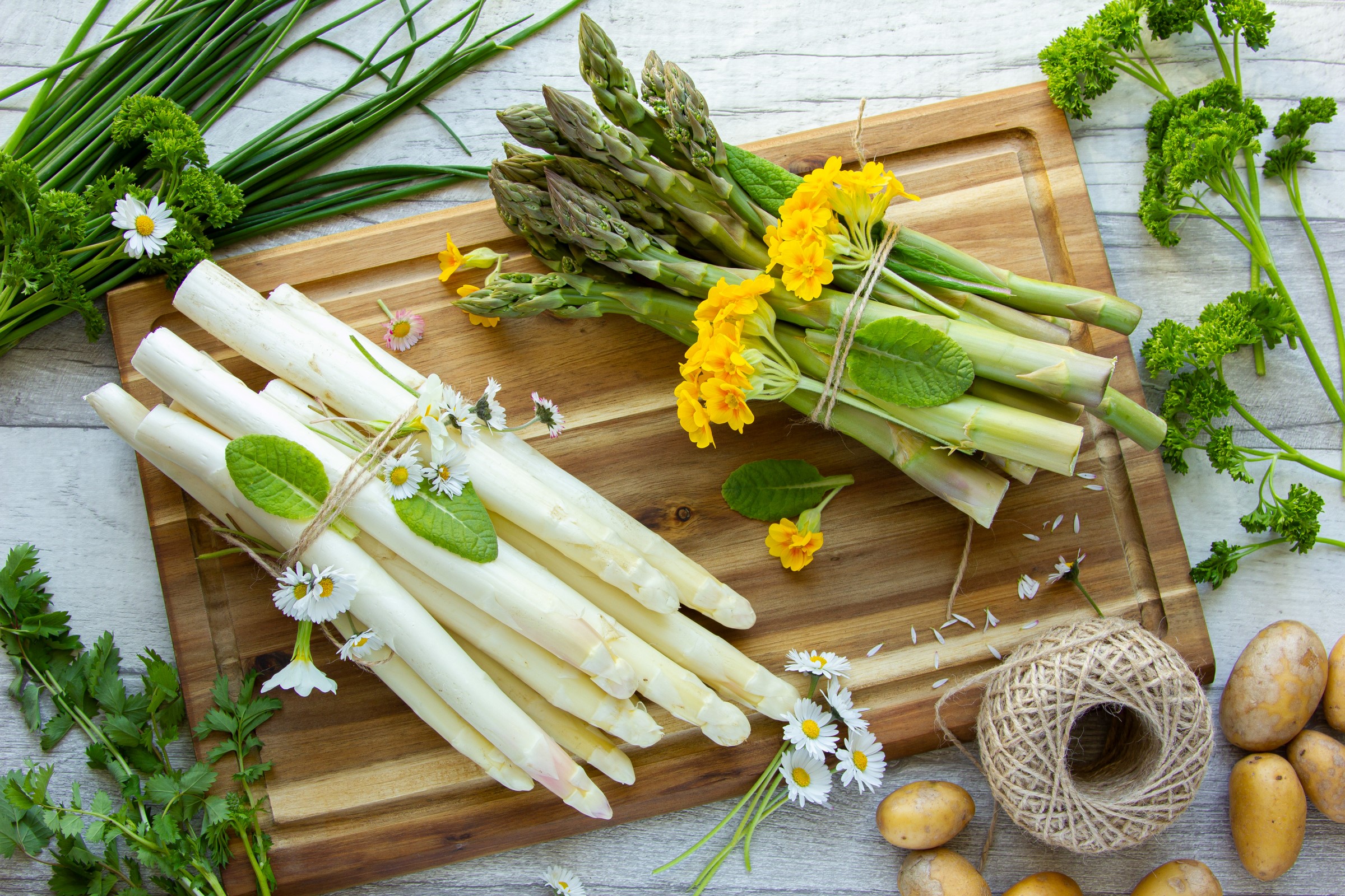Svazky bílého a zeleného chřestu s květy na dřevěném prkénku, obklopené bylinkami a bramborami.