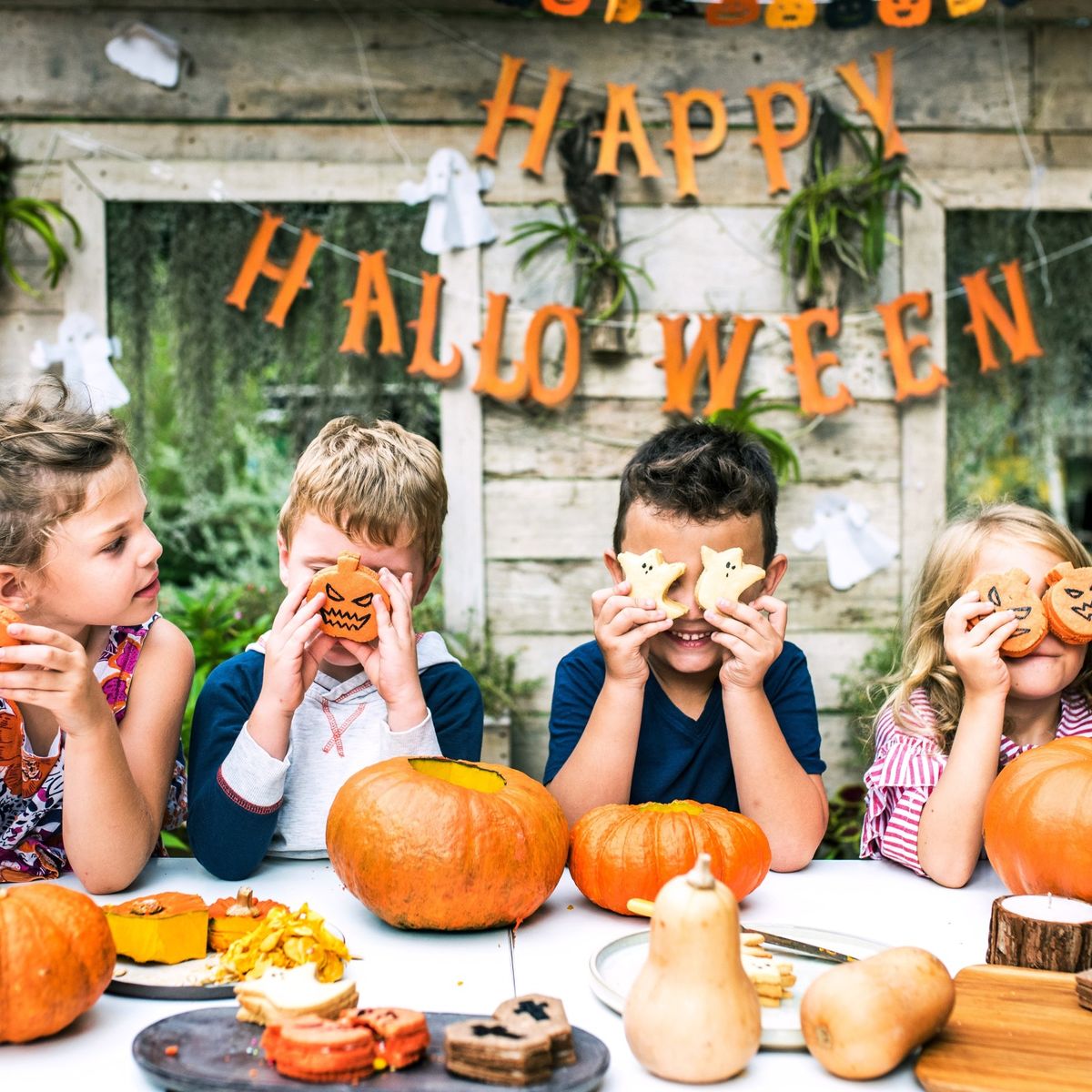 Děti sedí u stolu s dýněmi a drží si halloweenské sušenky u obličeje. Za nimi visí nápis „Happy Halloween“.
