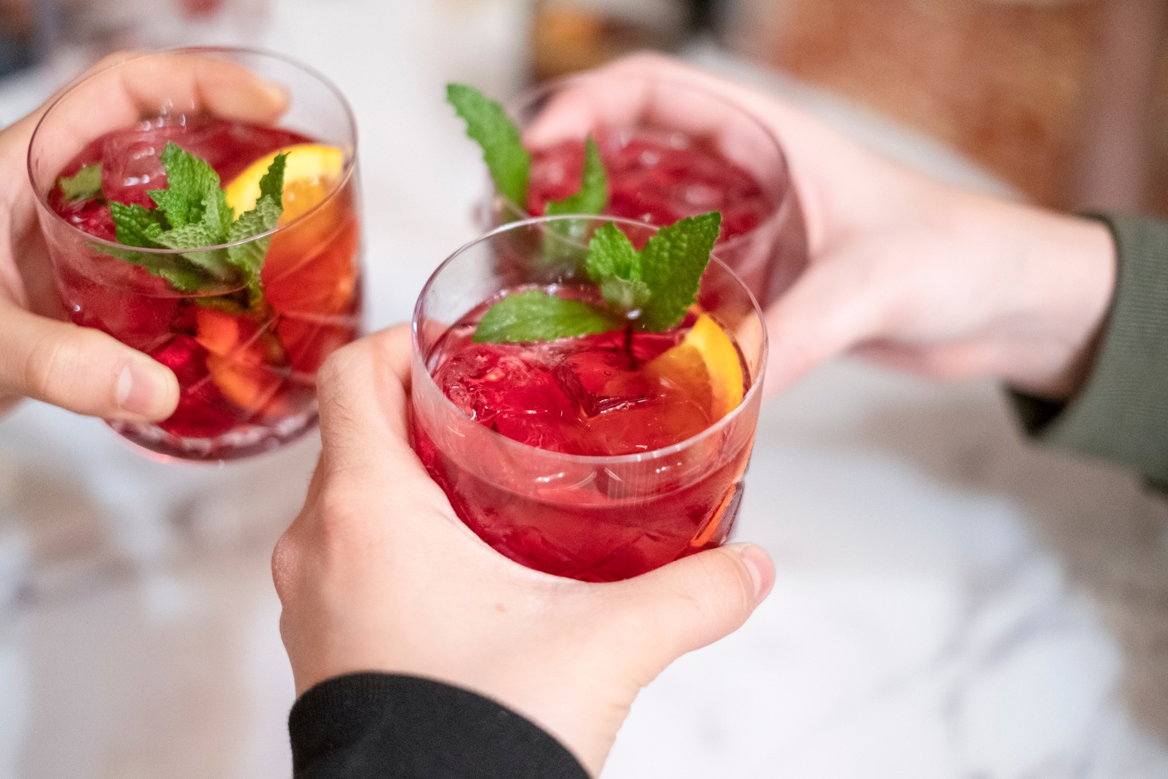 Tři ruce si na bílém pozadí připíjejí sklenicemi naplněnými sangrií, ozdobenými lístky máty a plátky pomeranče.