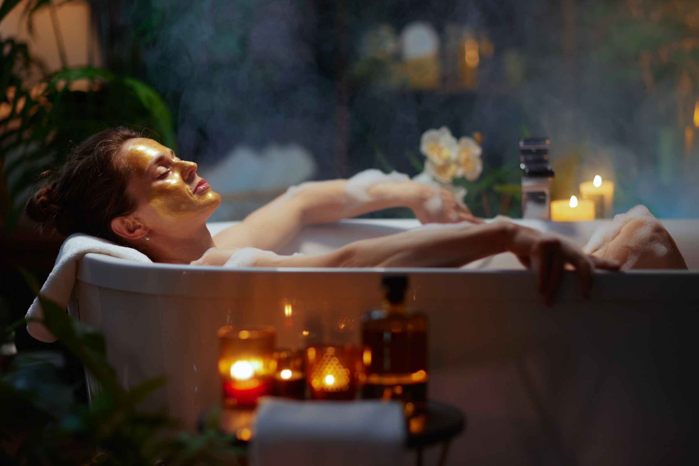Žena odpočívající v bublinkové koupeli se zlatou pleťovou maskou, obklopená svíčkami a rostlinami, což vytváří klidnou a útulnou atmosféru.