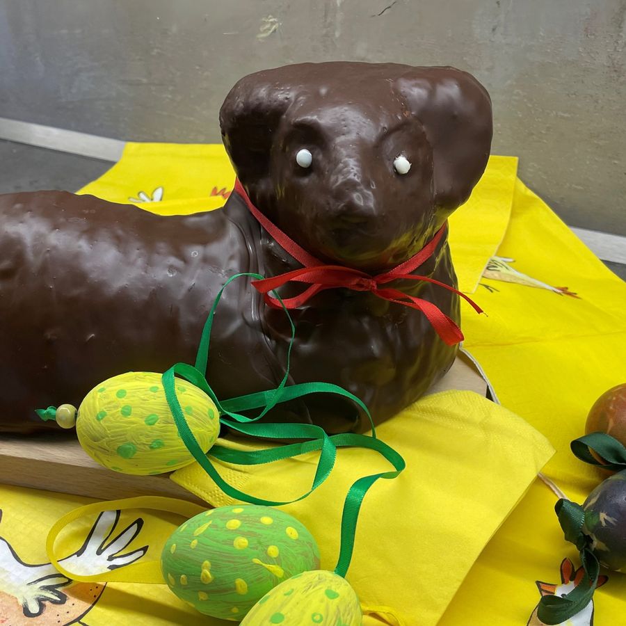 Čokoládový beránek s červenou stužkou na žlutém ubrusu, obklopený malovanými velikonočními vajíčky a ubrousky s motivy kuřátek.