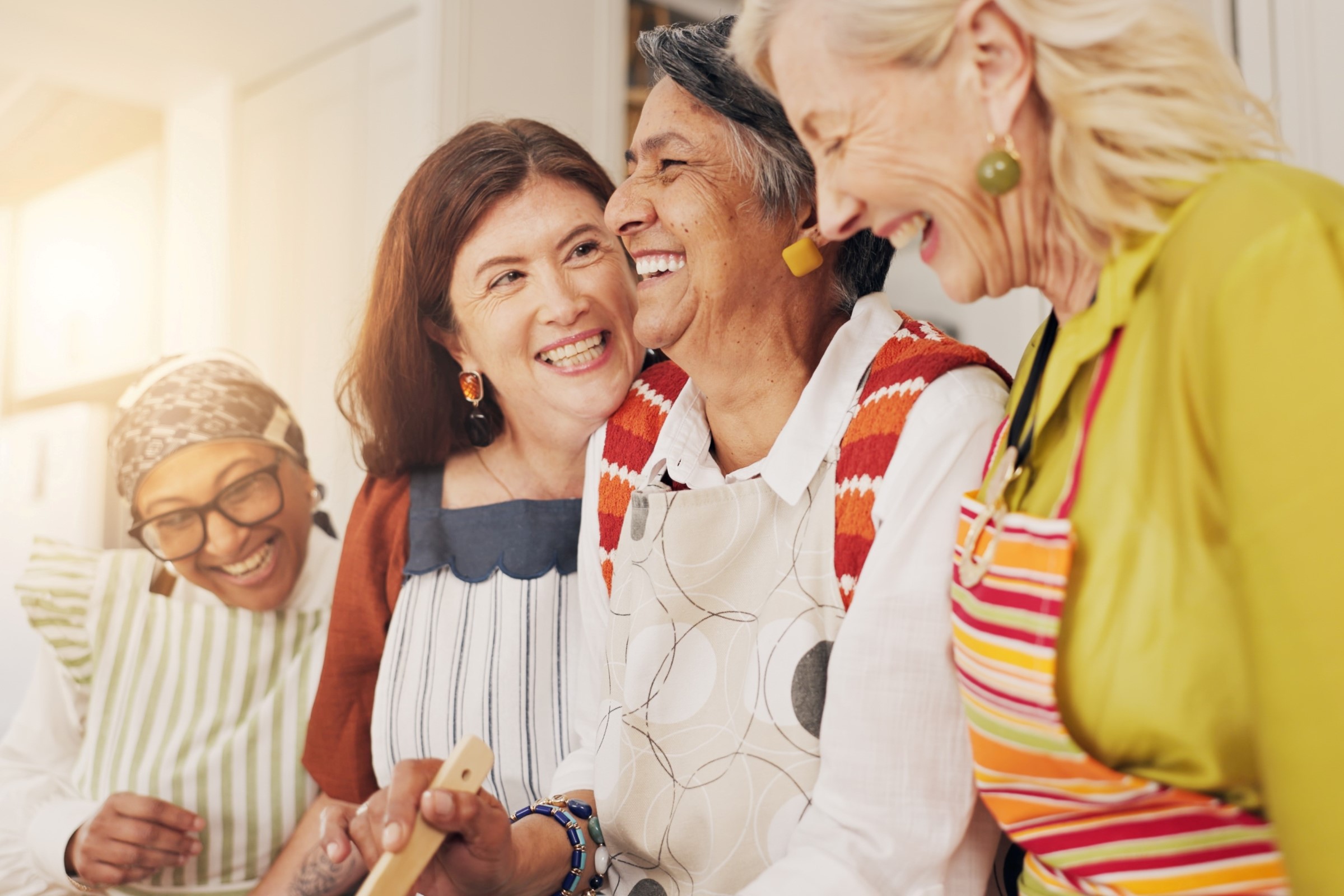 Čtyři ženy v zástěrách se smějí a užívají si společné chvíle ve světlé kuchyni.