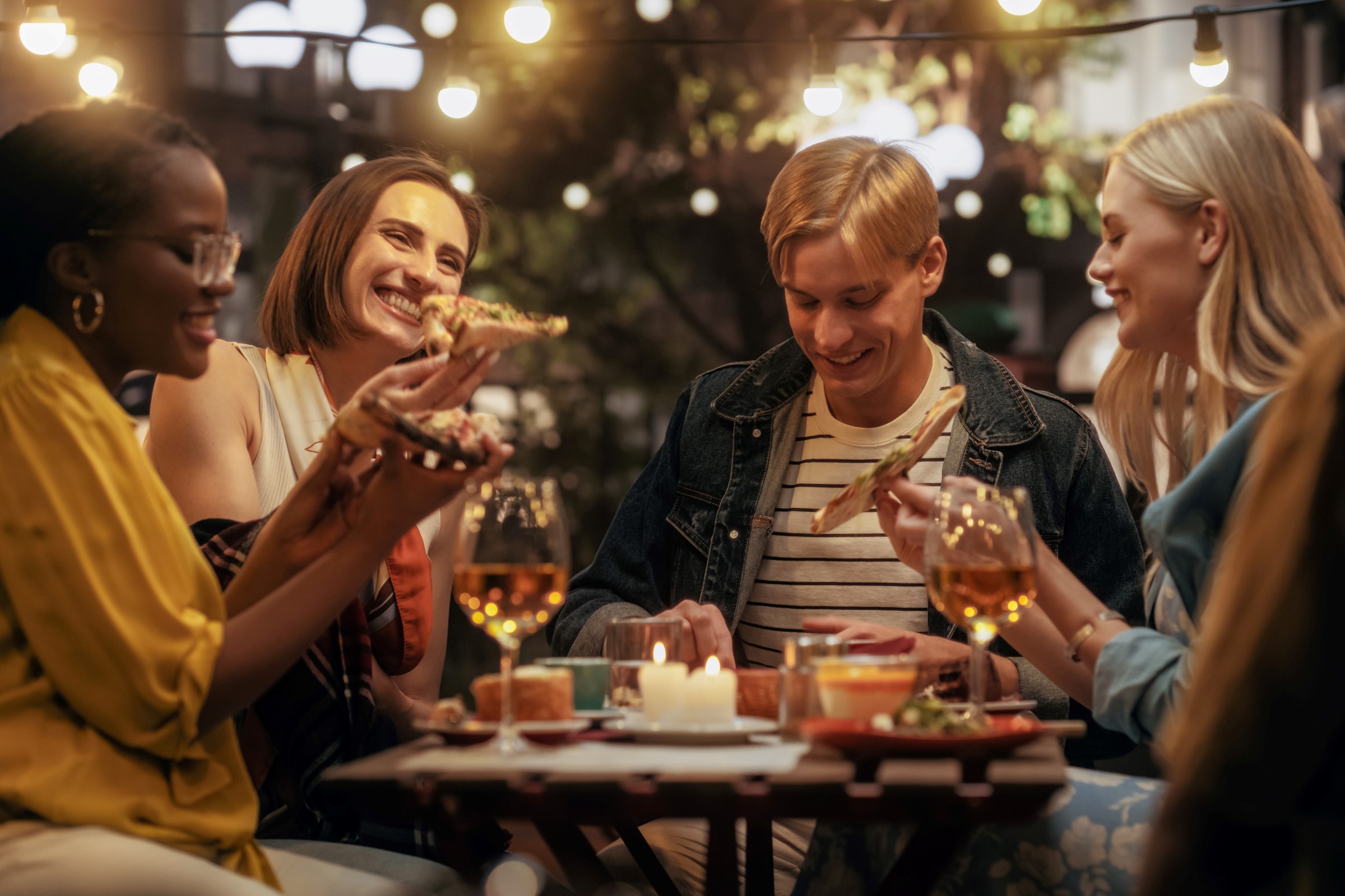 Čtyři přátelé si s radostí pochutnávají na pizze a popíjejí drinky v útulné venkovní restauraci osvětlené řetězovými světýlky.