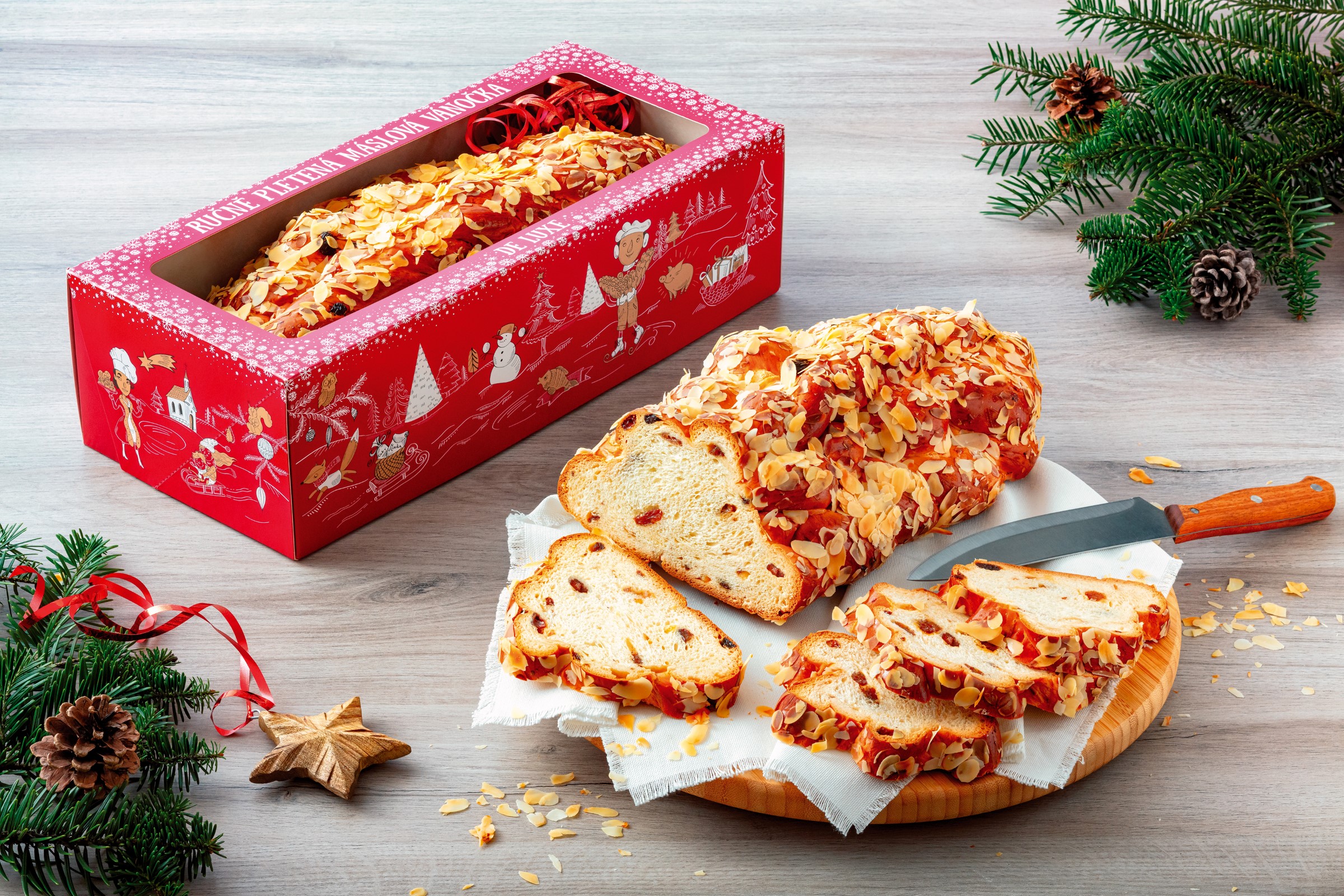 Nakrájená Máslová vánočka de luxe Globus posypaná mandlemi na dřevěné desce, vedle ní slavnostní červená krabička s vánočkou uvnitř, obklopená borovicovými větvemi a šiškami.
