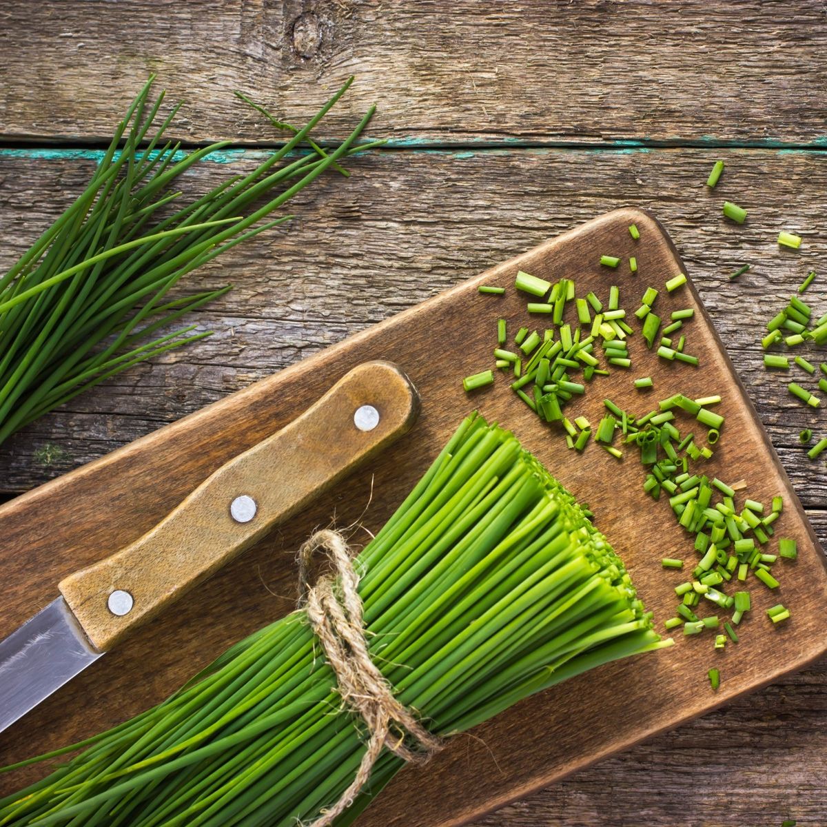 Čerstvá pažitka svázaná provázkem na dřevěné prkénko, s nožem a nasekanou pažitkou rozsypanou poblíž na rustikálním dřevěném stole.