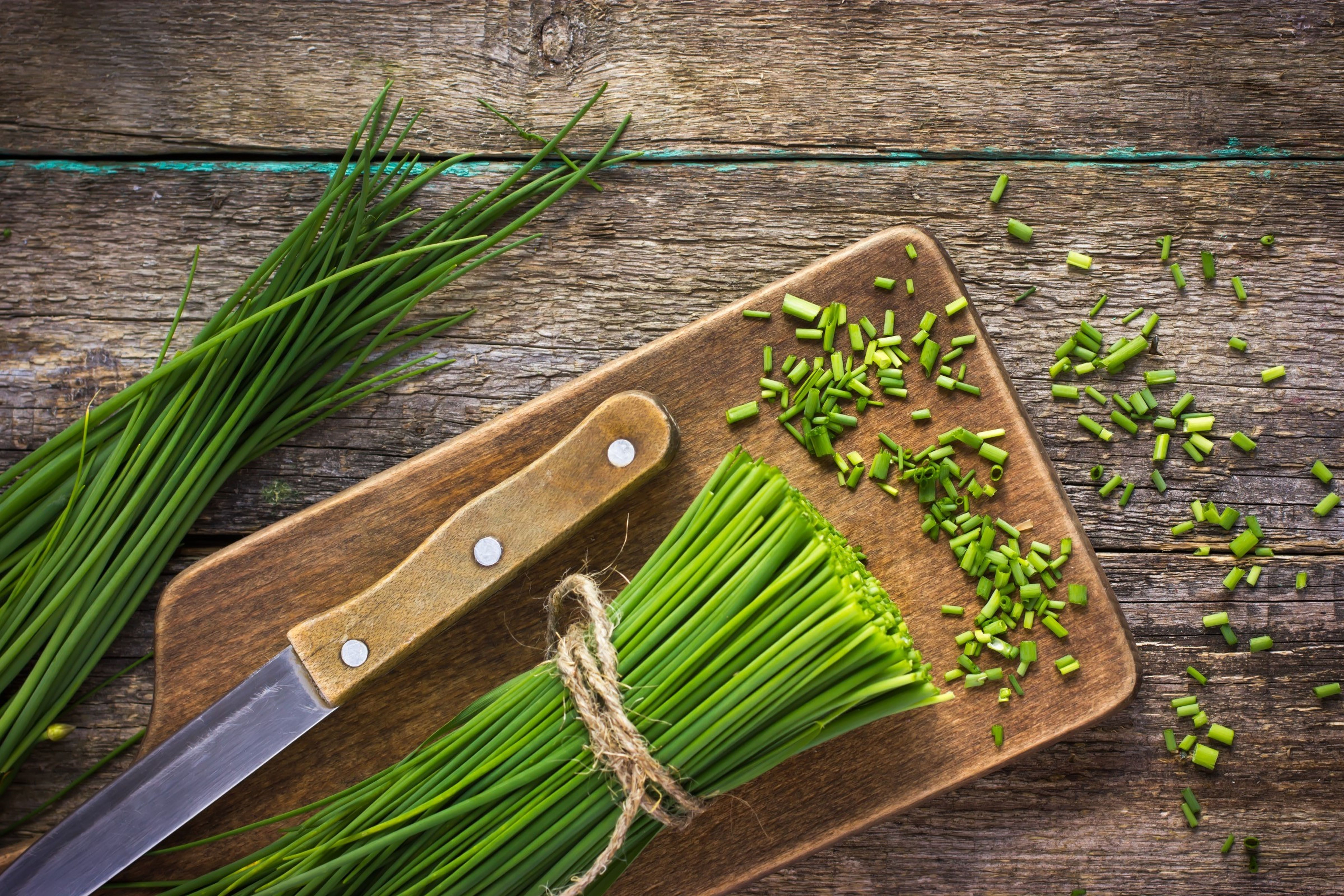 Čerstvá pažitka svázaná provázkem na dřevěné prkénko, s nožem a nasekanou pažitkou rozsypanou poblíž na rustikálním dřevěném stole.