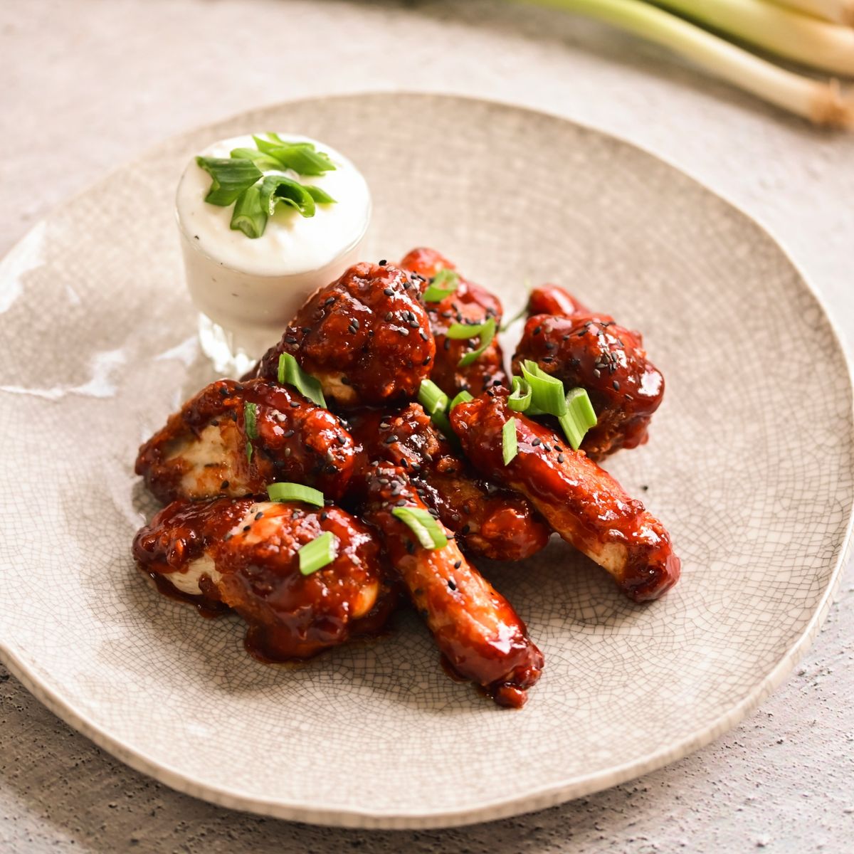 Kuřecí křidélka v BBQ omáčce ozdobená jarní cibulkou, podávaná s mističkou krémového dipu na strukturovaném talíři. Recept připravil Vojta Poštulka.