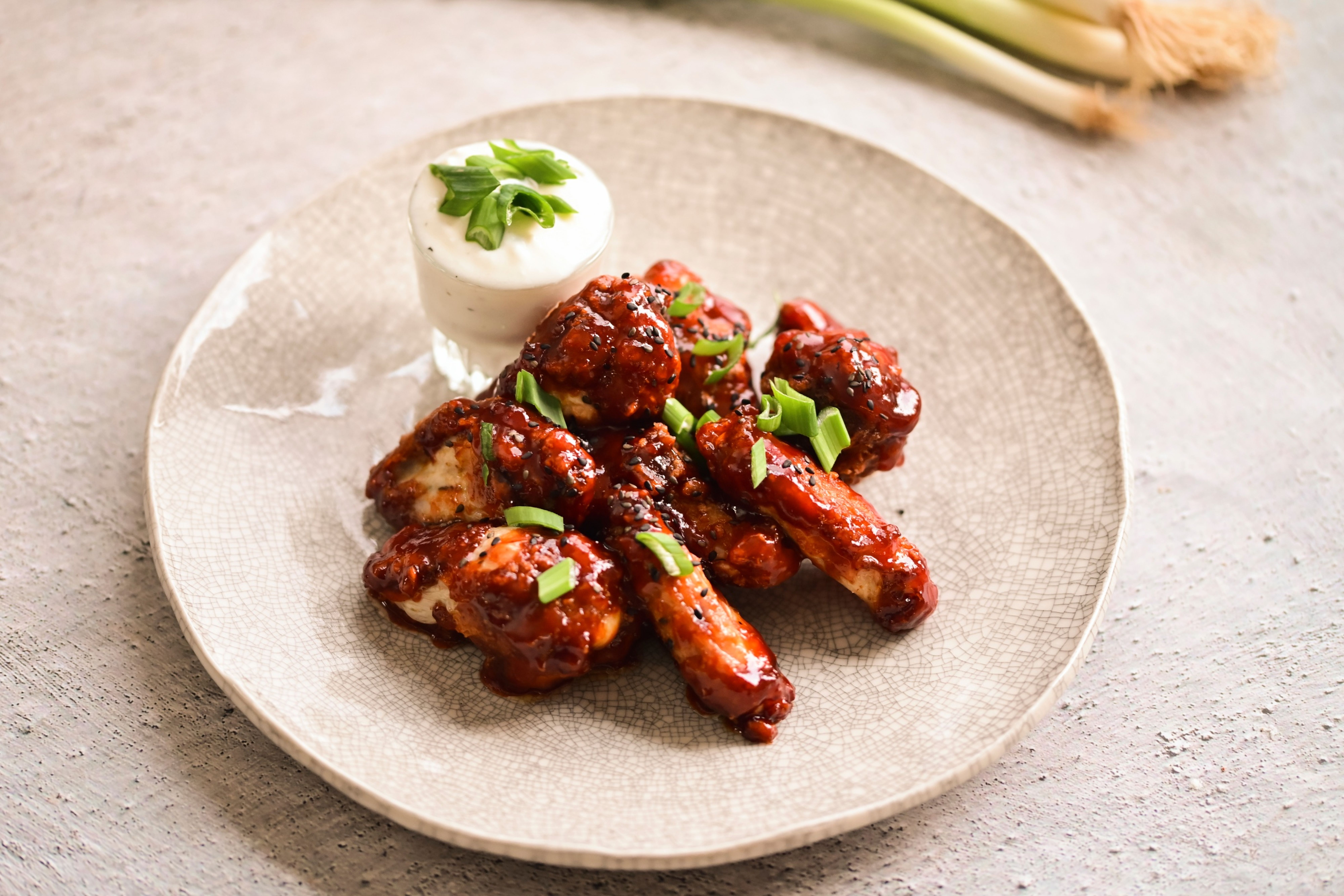 Kuřecí křidélka v BBQ omáčce ozdobená jarní cibulkou, podávaná s mističkou krémového dipu na strukturovaném talíři. Recept připravil Vojta Poštulka.