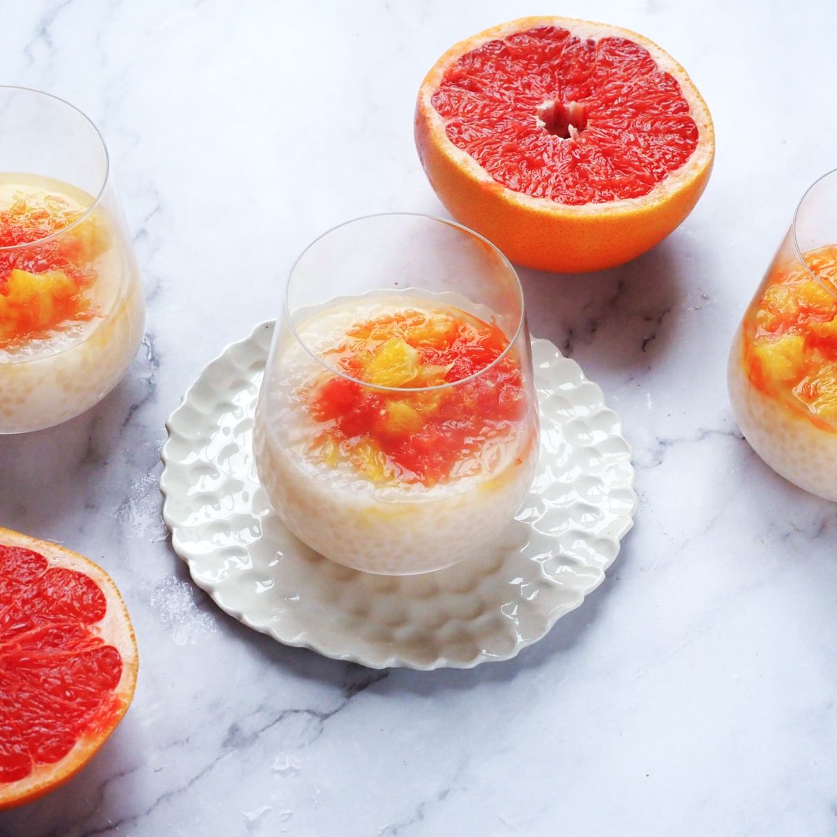 Tři sklenice krémového tapiokového pudinku s grepovým kompotem, obklopené dvěma rozpůlenými růžovými grapefruity na mramorovém povrchu.