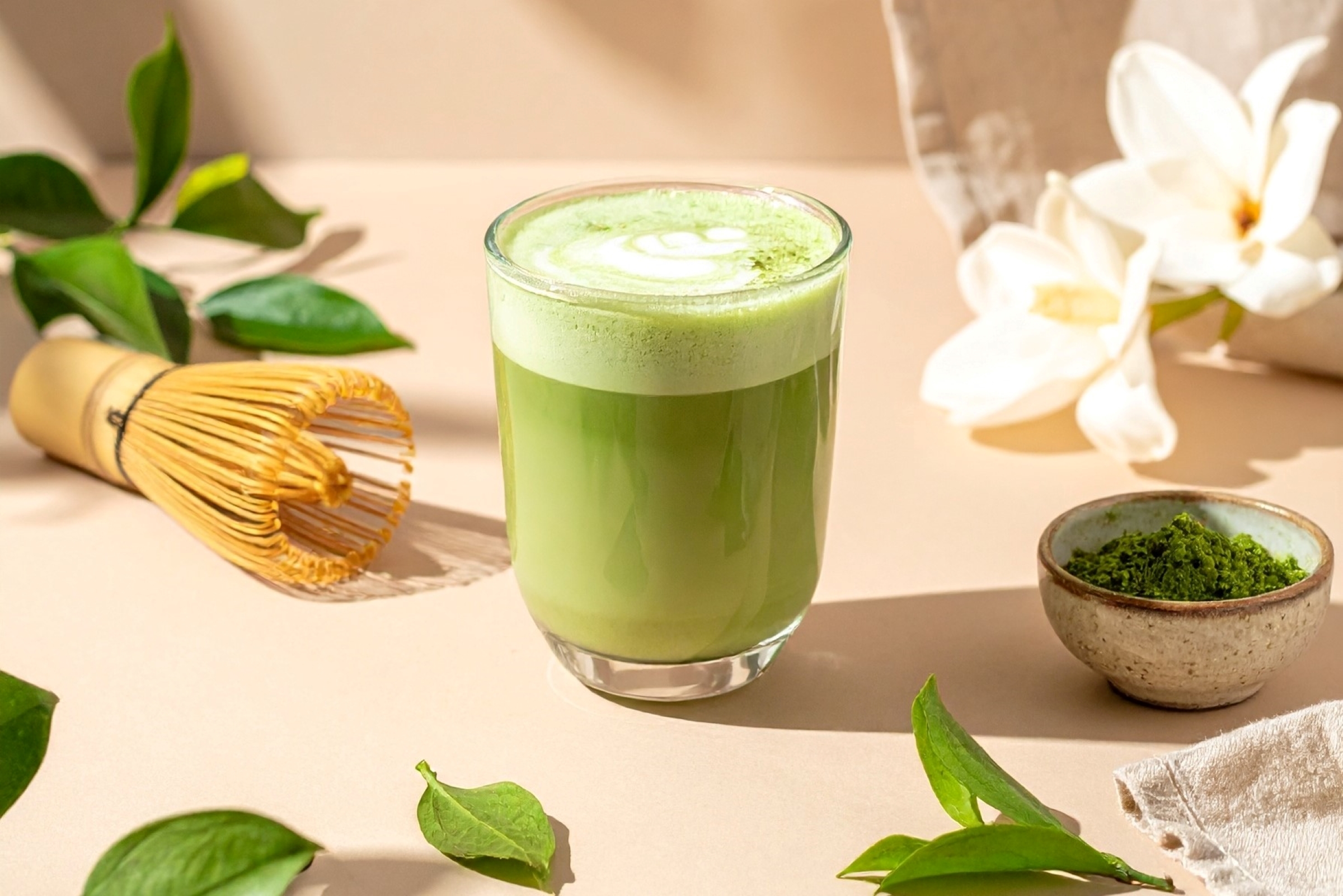 Sklenice napěněného matcha latte vedle bambusové metličky, prášek matcha v misce, zelené lístky a bílé květy na světlém podkladu.