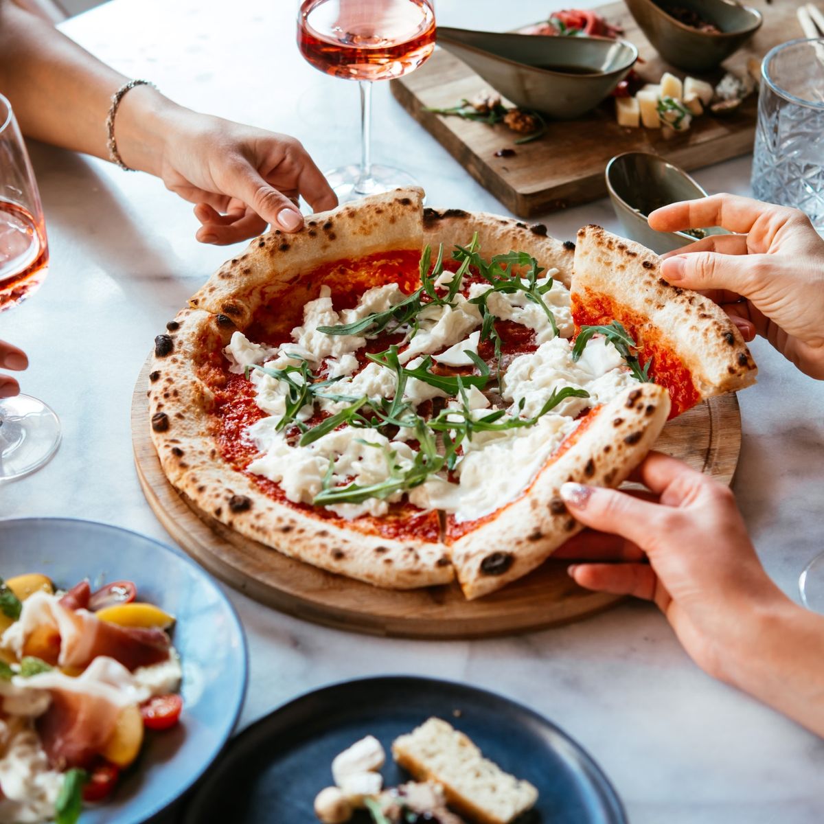 Tři lidé, kteří si u stolu s růžovým vínem a různými předkrmy dělí pizzu s rukolou a mozzarellou.