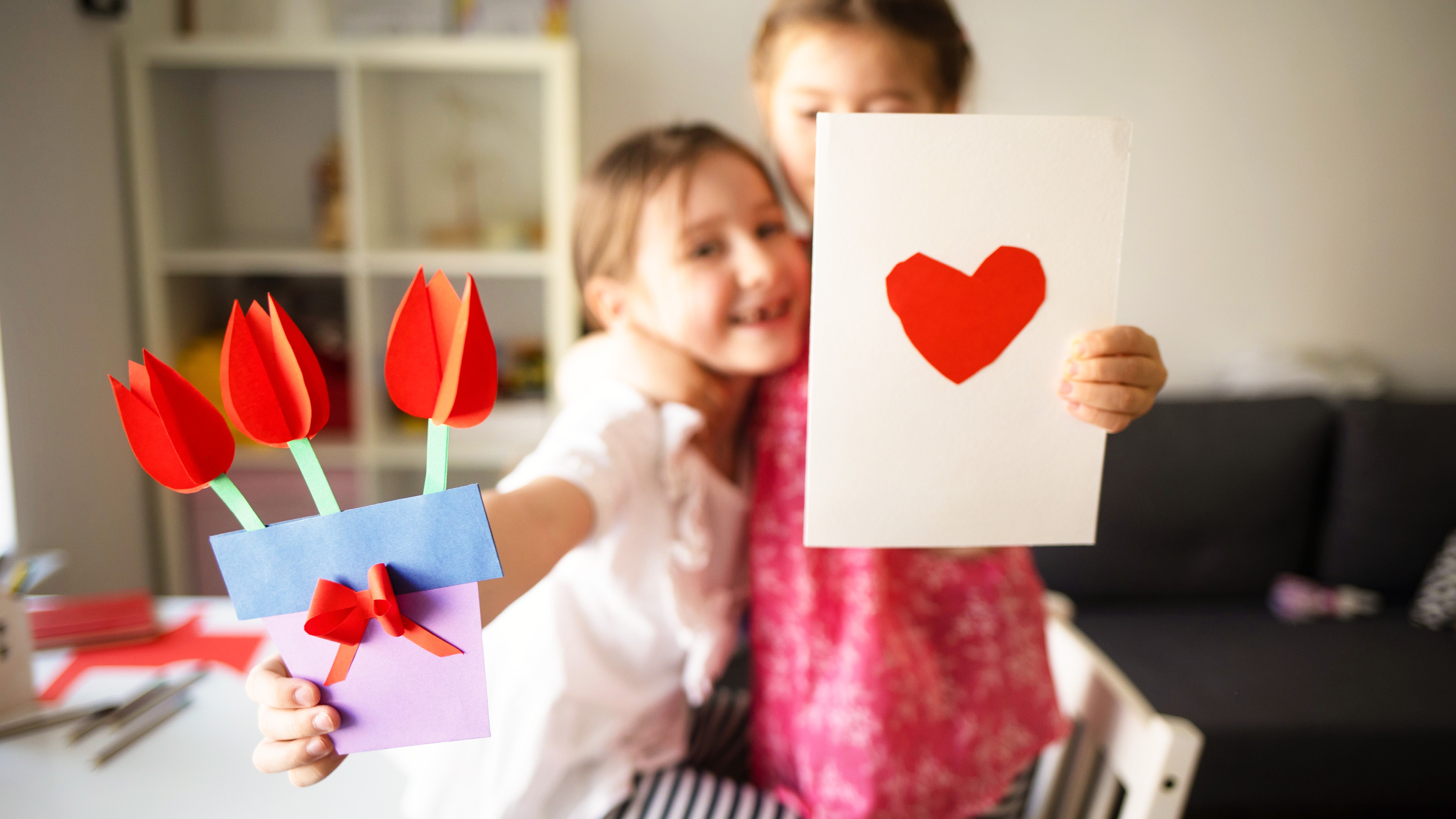 Dvě děti se usmívají a drží papírové přání s červeným srdcem a kyticí papírových tulipánů.