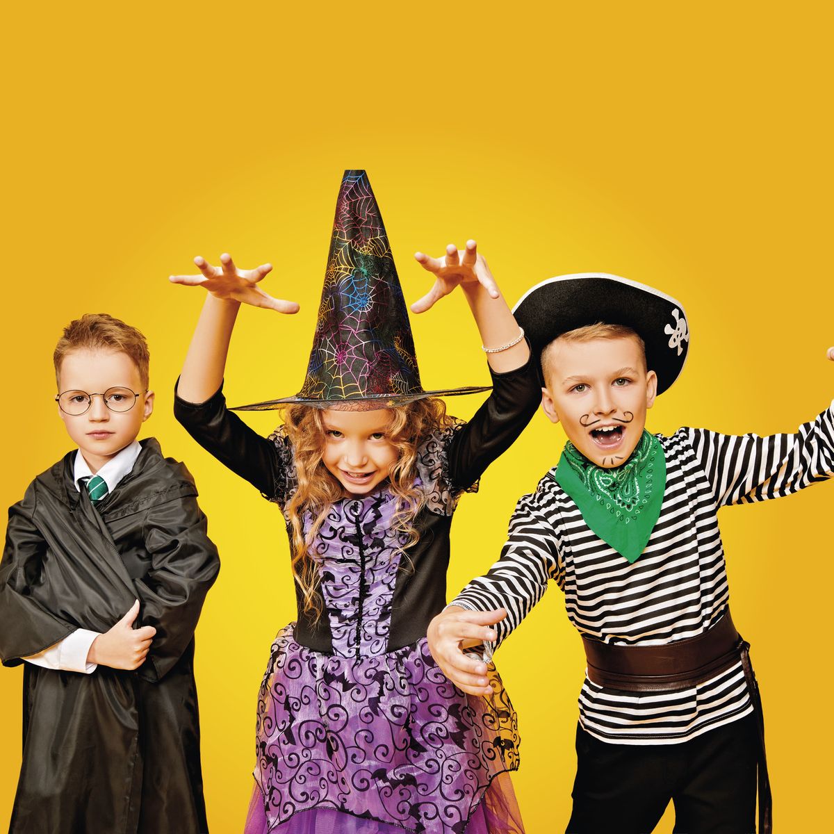 Na žlutém pozadí stojí tři děti v kostýmech: jedno v čarodějnickém rouchu, jedno v čarodějnickém oblečení a jedno v kostýmu piráta.